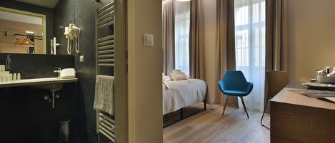 A modern hotel room with a separate bathroom, a bed with a white duvet, a blue chair, and a desk with a lamp.