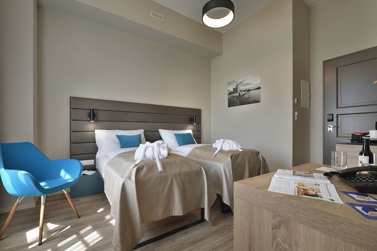 A hotel room with two beds, a blue chair, a wooden desk, and a wall-mounted picture.