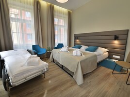 Quarto de hotel com duas camas, uma mesinha de cabeceira, uma cadeira e uma janela grande com cortinas.