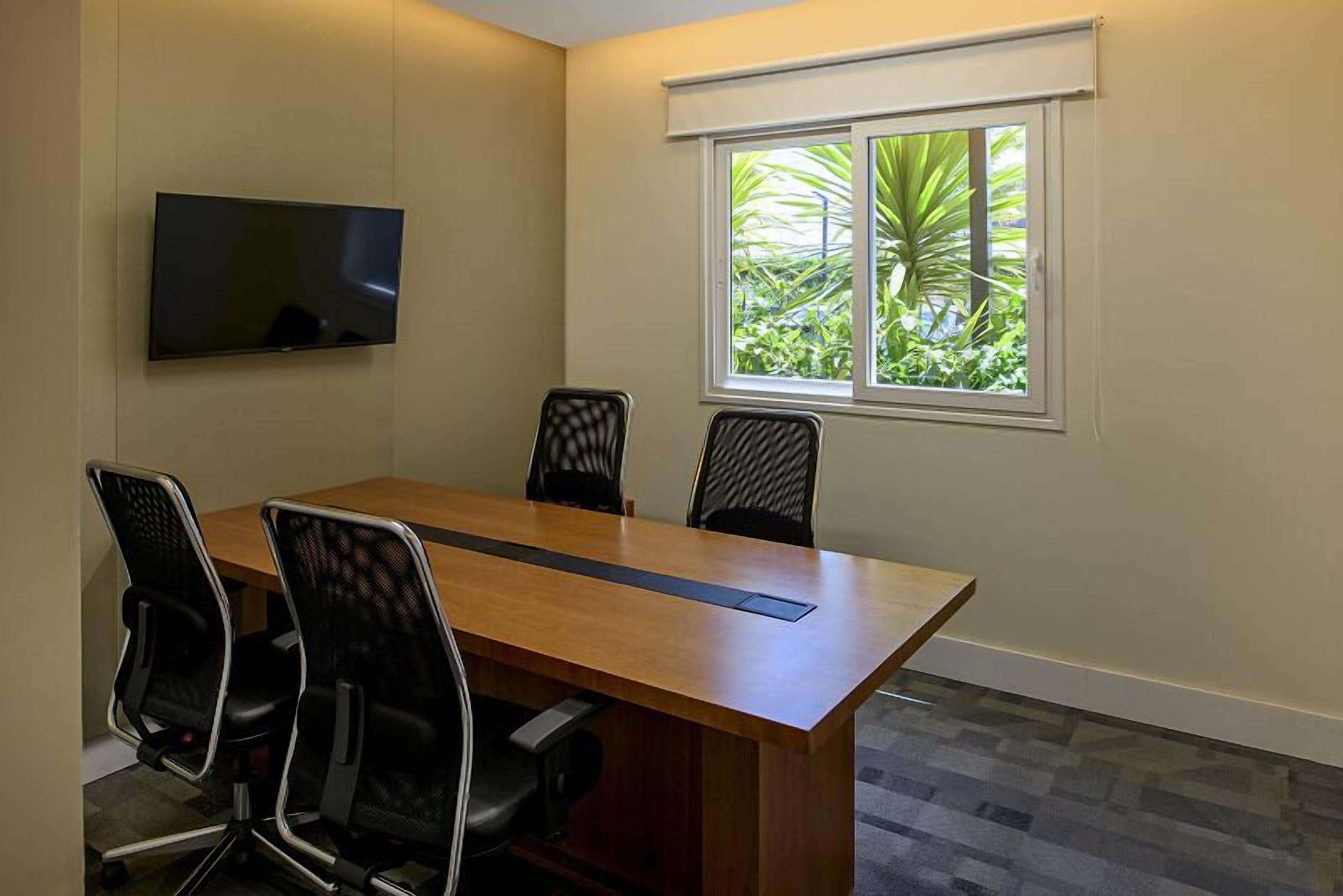 Uma sala de conferência com uma mesa comprida, quatro cadeiras, uma televisão de tela plana e uma janela com vista para o verde.