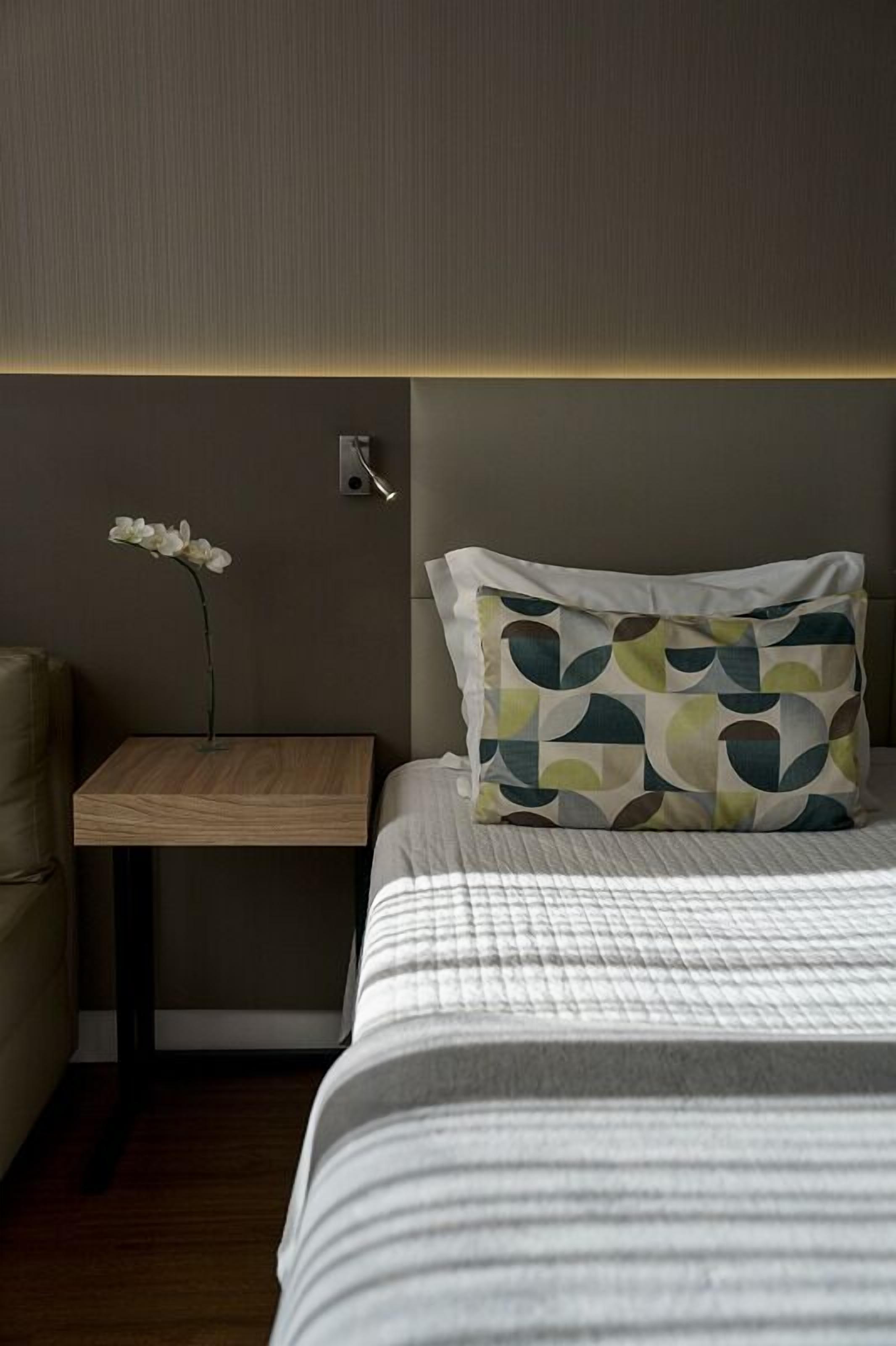 A neatly made bed with a patterned pillow, a small bedside table with a vase of flowers, and a wall-mounted light fixture.