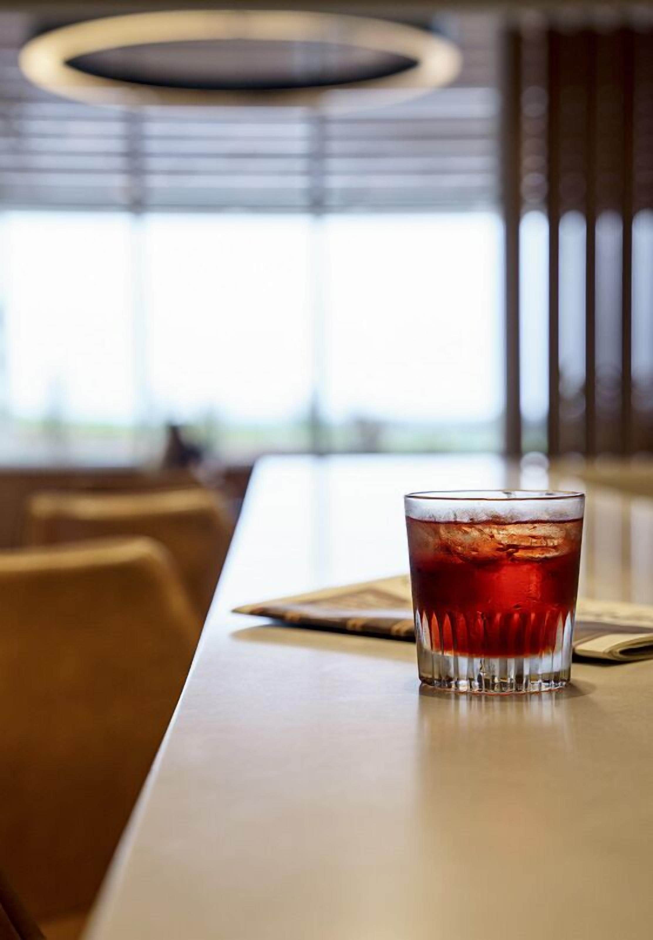 A glass of red beverage on a table in a restaurant with a view of the outdoors.