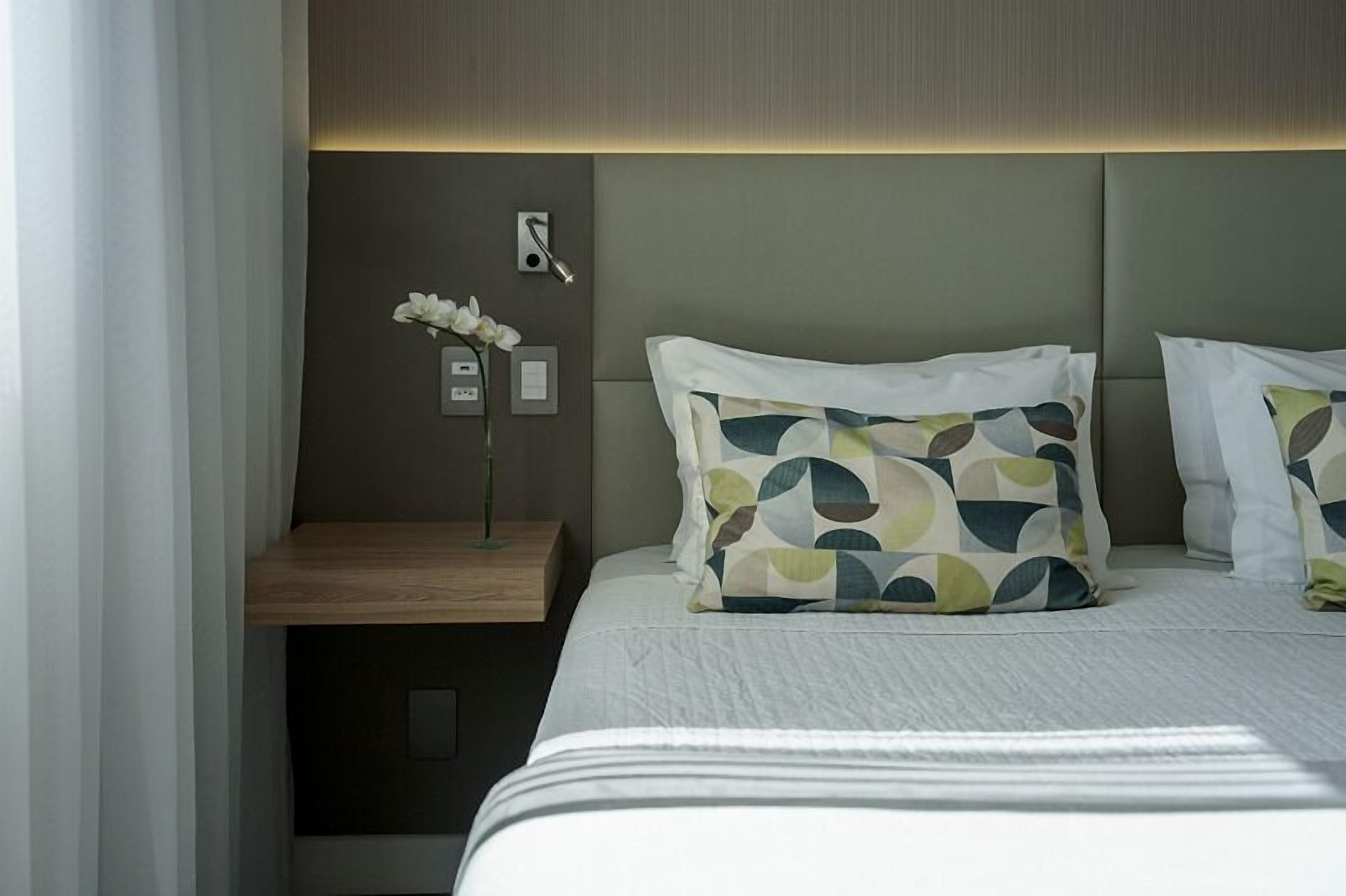 A hotel room with a bed, a bedside table with a vase of flowers, and a wall-mounted control panel.