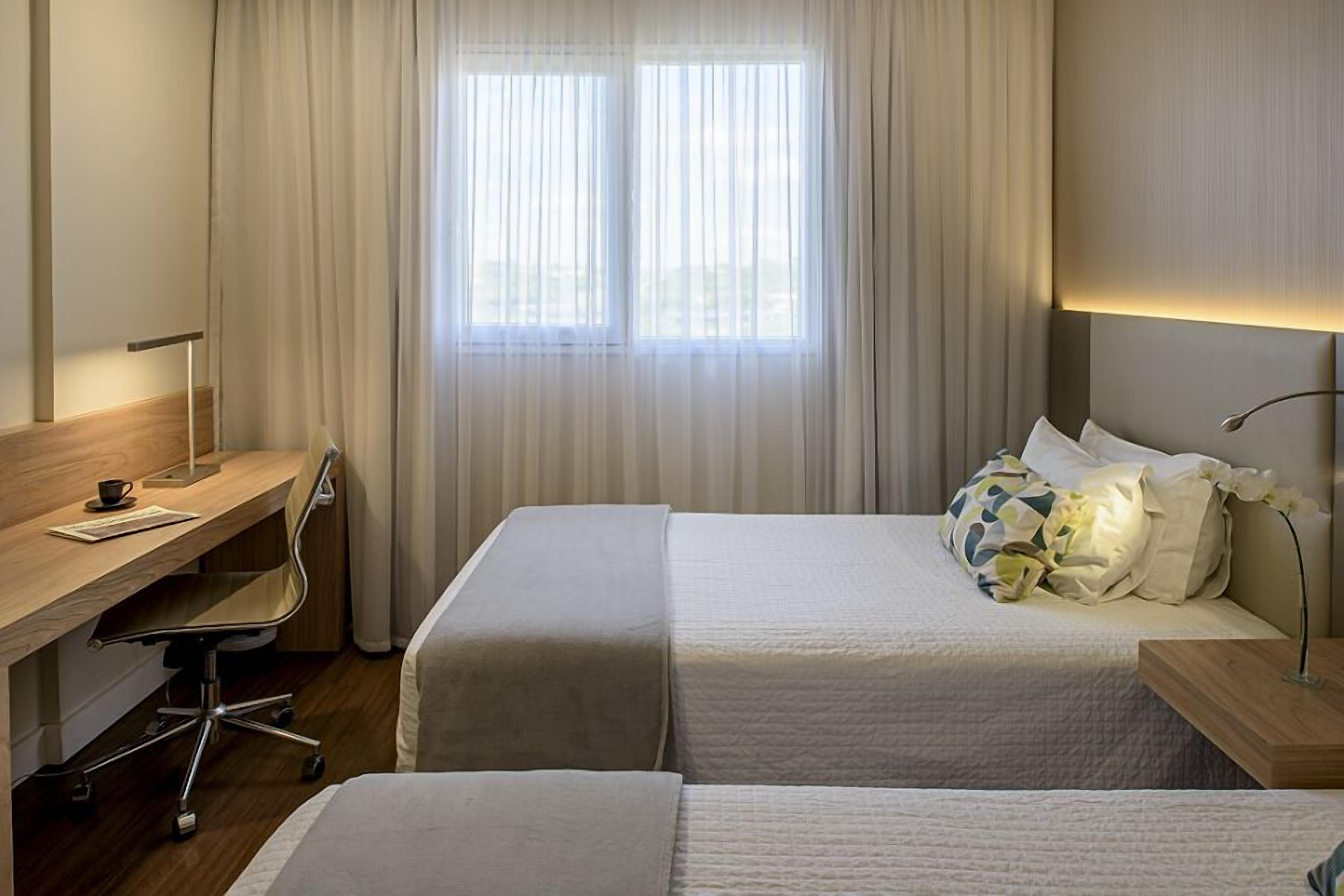 A hotel room with two beds, a desk, a chair, and a window with curtains.