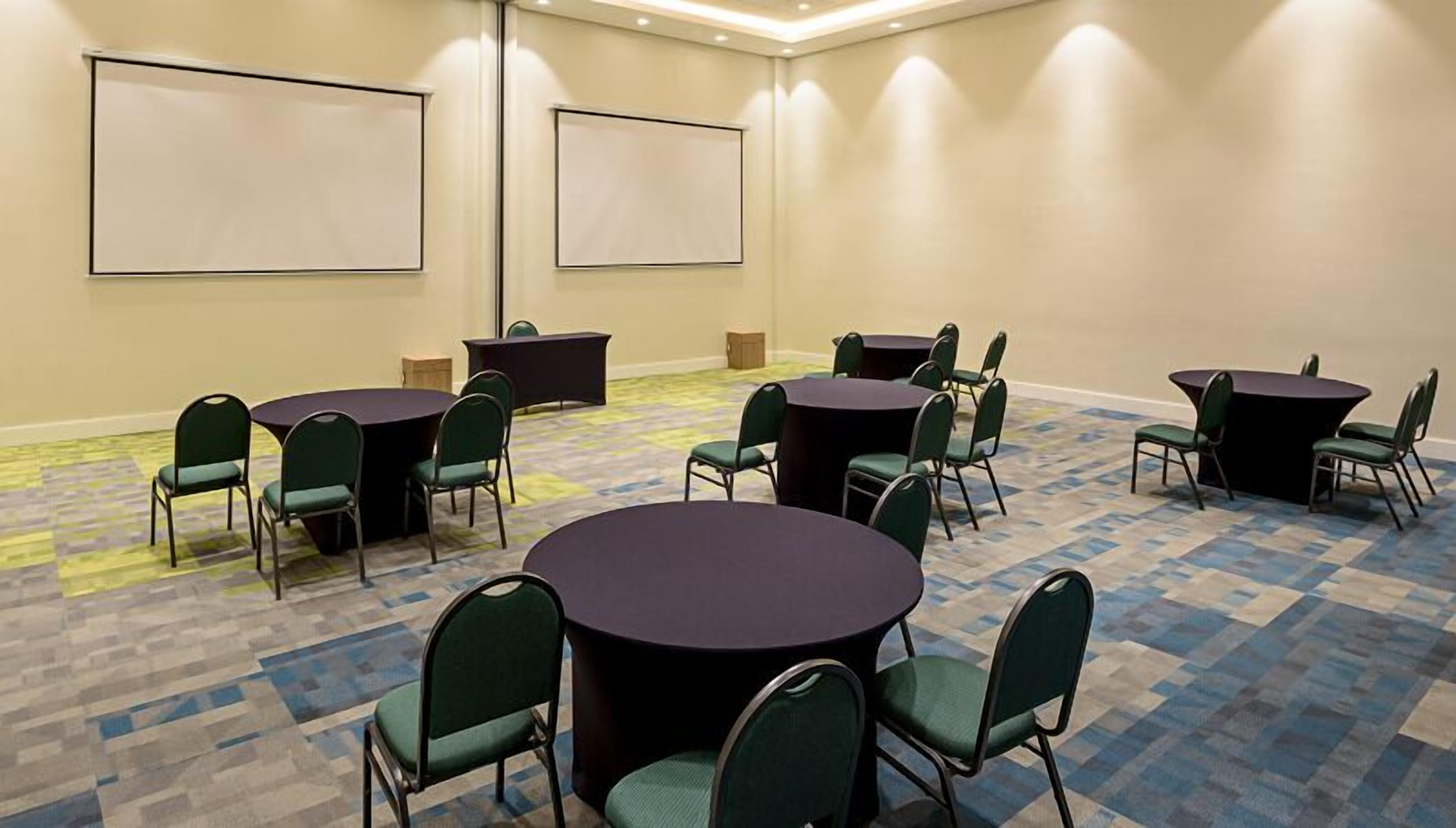 A conference room with multiple tables and chairs, a projector screen, and carpeted flooring.