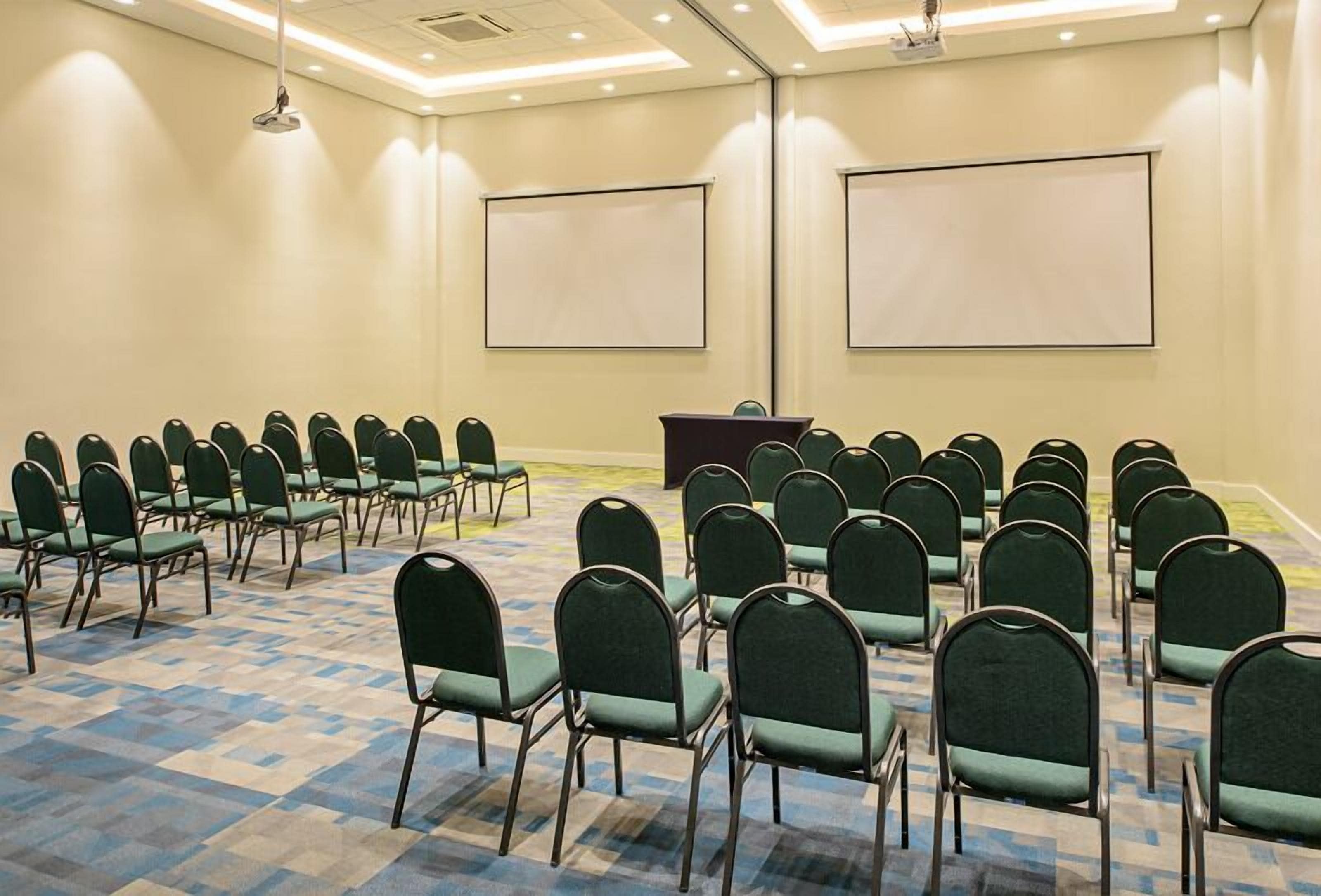 Un salón de conferencias con filas de sillas verdes, un atril y dos pantallas de proyección.