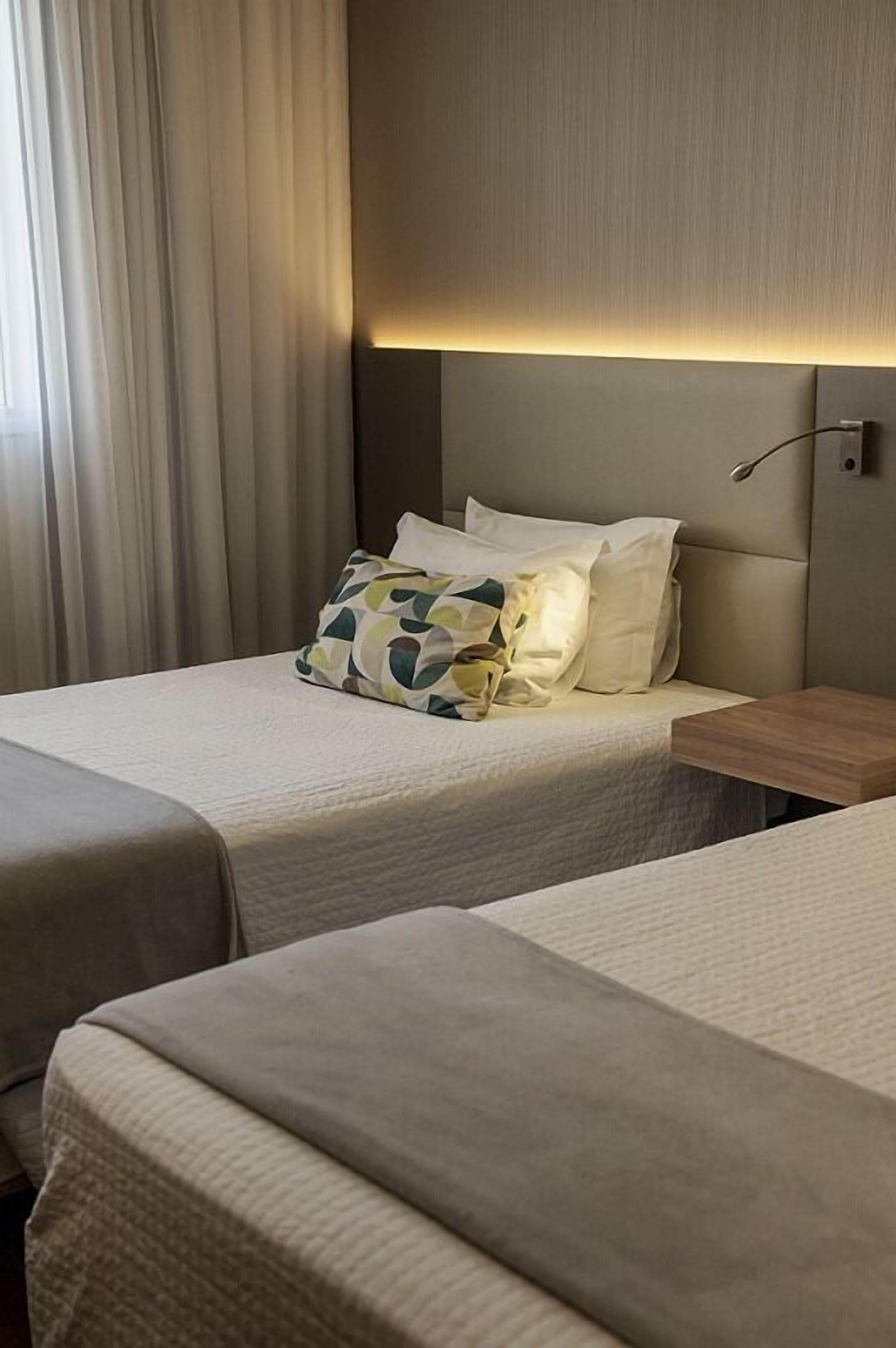 A hotel room with two beds, a headboard with integrated lighting, a bedside table, and a window with curtains.