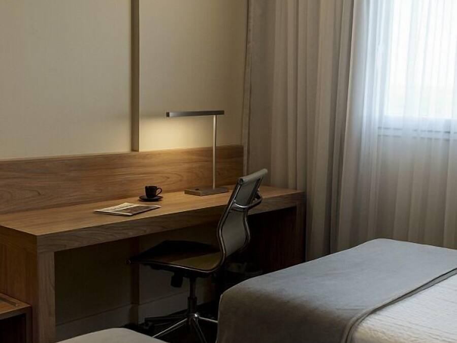 A hotel room with a wooden desk, a chair, two beds, and a window with curtains.