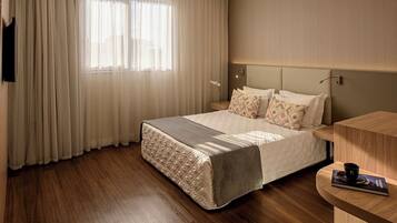 Quarto de hotel com cama, mesa de cabeceira, janela com cortinas e televisão na parede.