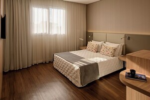 Quarto de hotel com cama, mesa de cabeceira, janela com cortinas e televisão na parede.