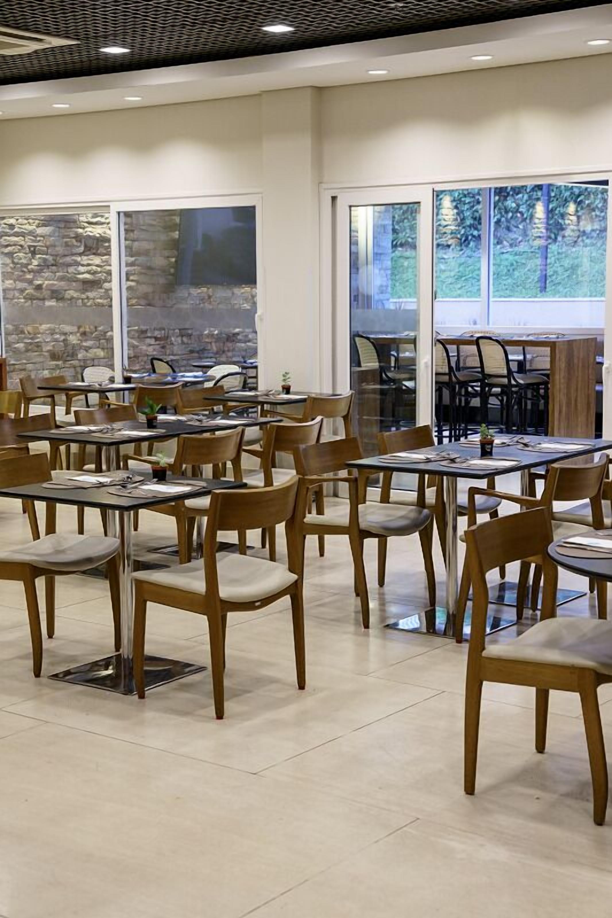 A spacious indoor dining area with multiple tables and chairs, large windows with a view of trees, and a stone wall.