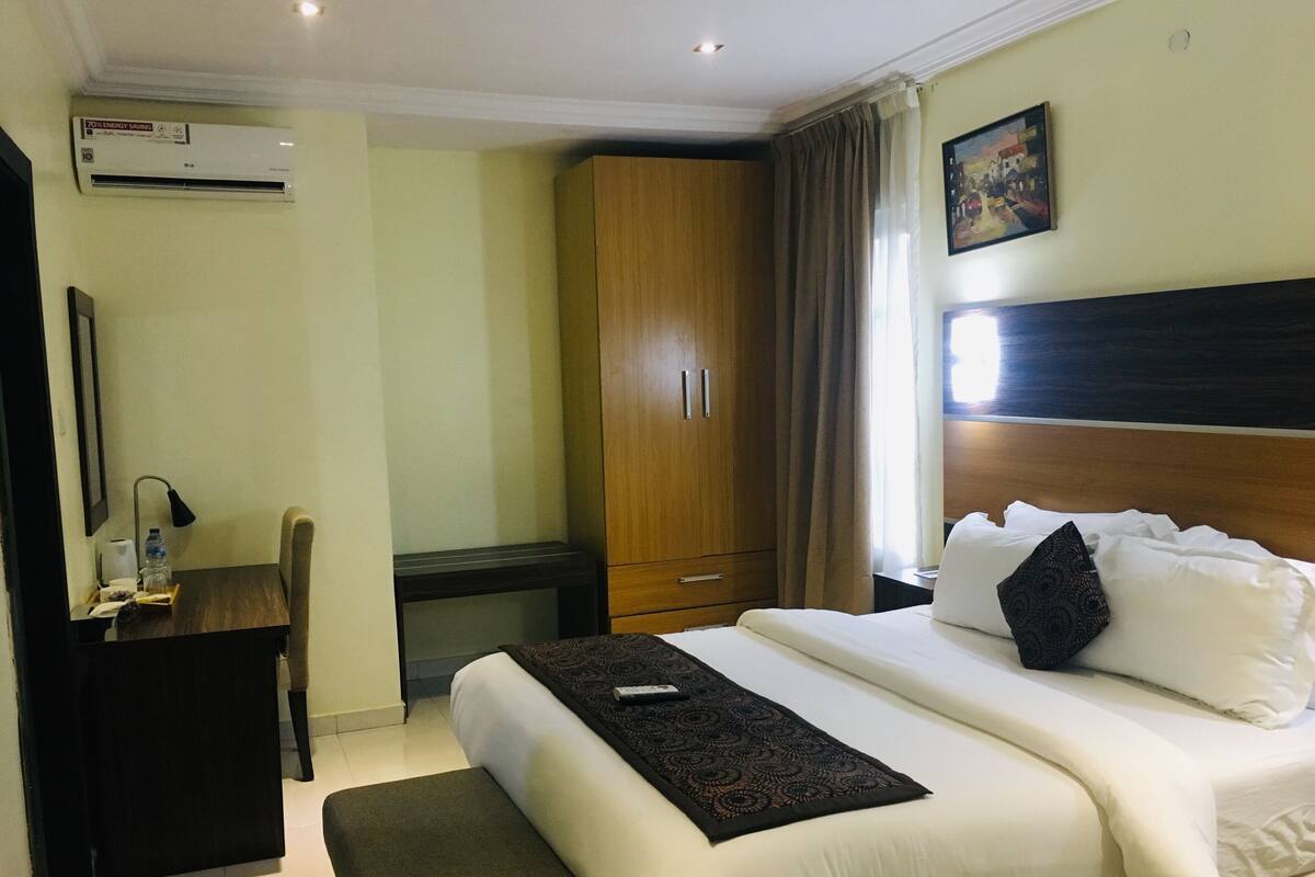 A hotel room with a bed, a desk, a chair, a wardrobe, and an air conditioning unit.