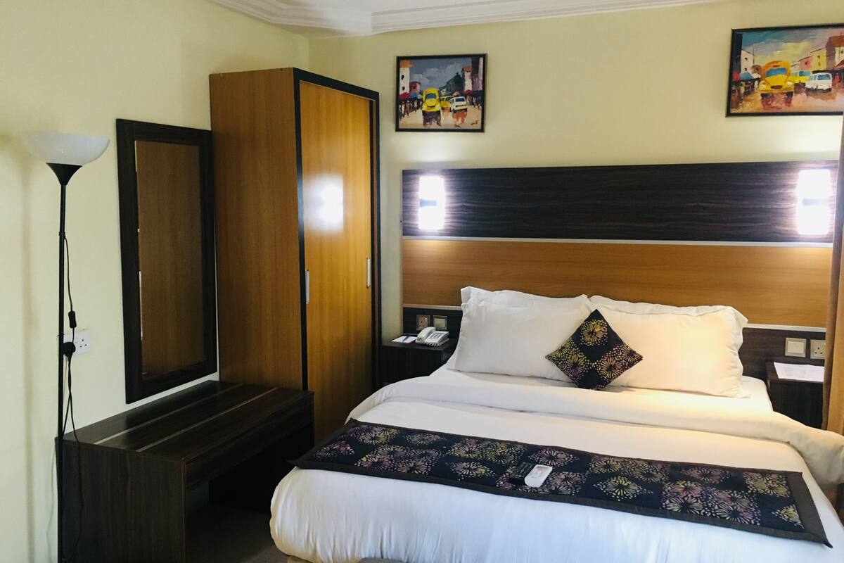 A hotel room with a large bed, a wardrobe, a bedside table with a lamp, and two paintings on the wall.