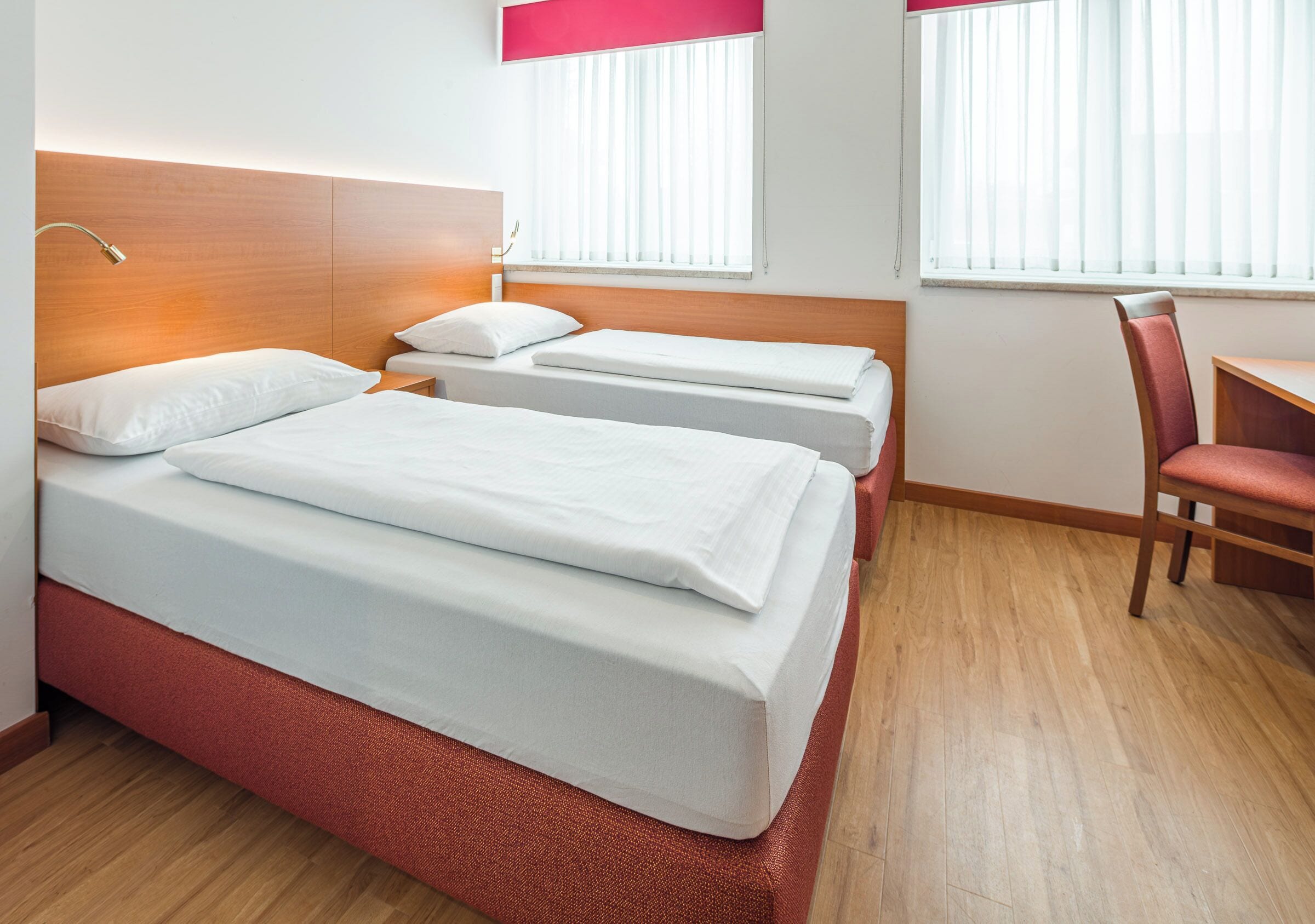 Une chambre d’hôtel avec une tête de lit en bois, deux lits simples, un bureau et une chaise.