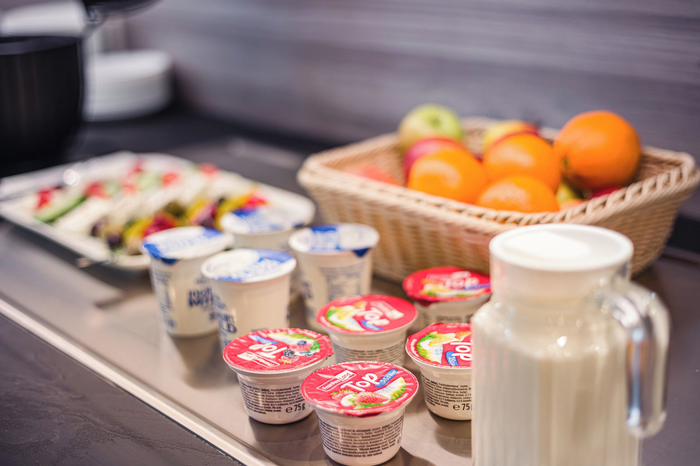 Una mesa de buffet con vasos de yogur, una cesta de fruta y una jarra de leche.