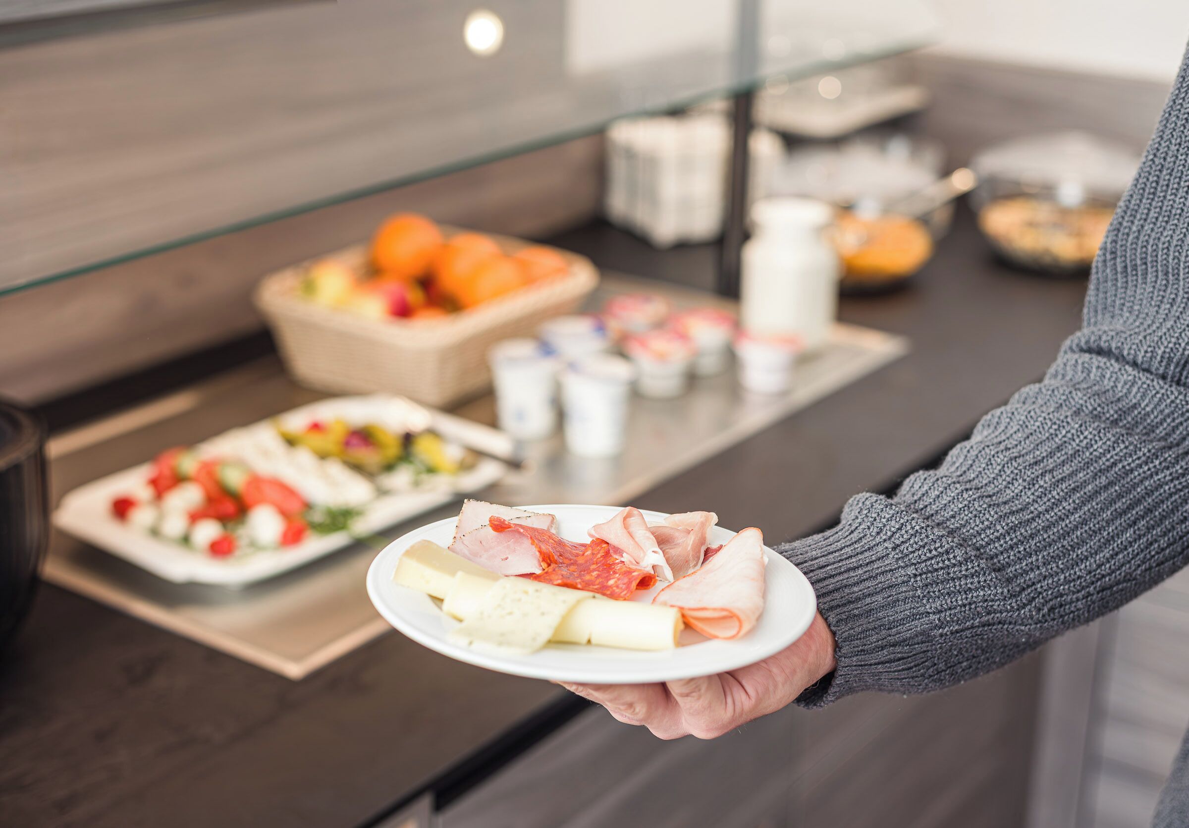 A person holding a plate with various food items, including cheese and cured meats, in a modern kitchen setting with a glass display case in the background.