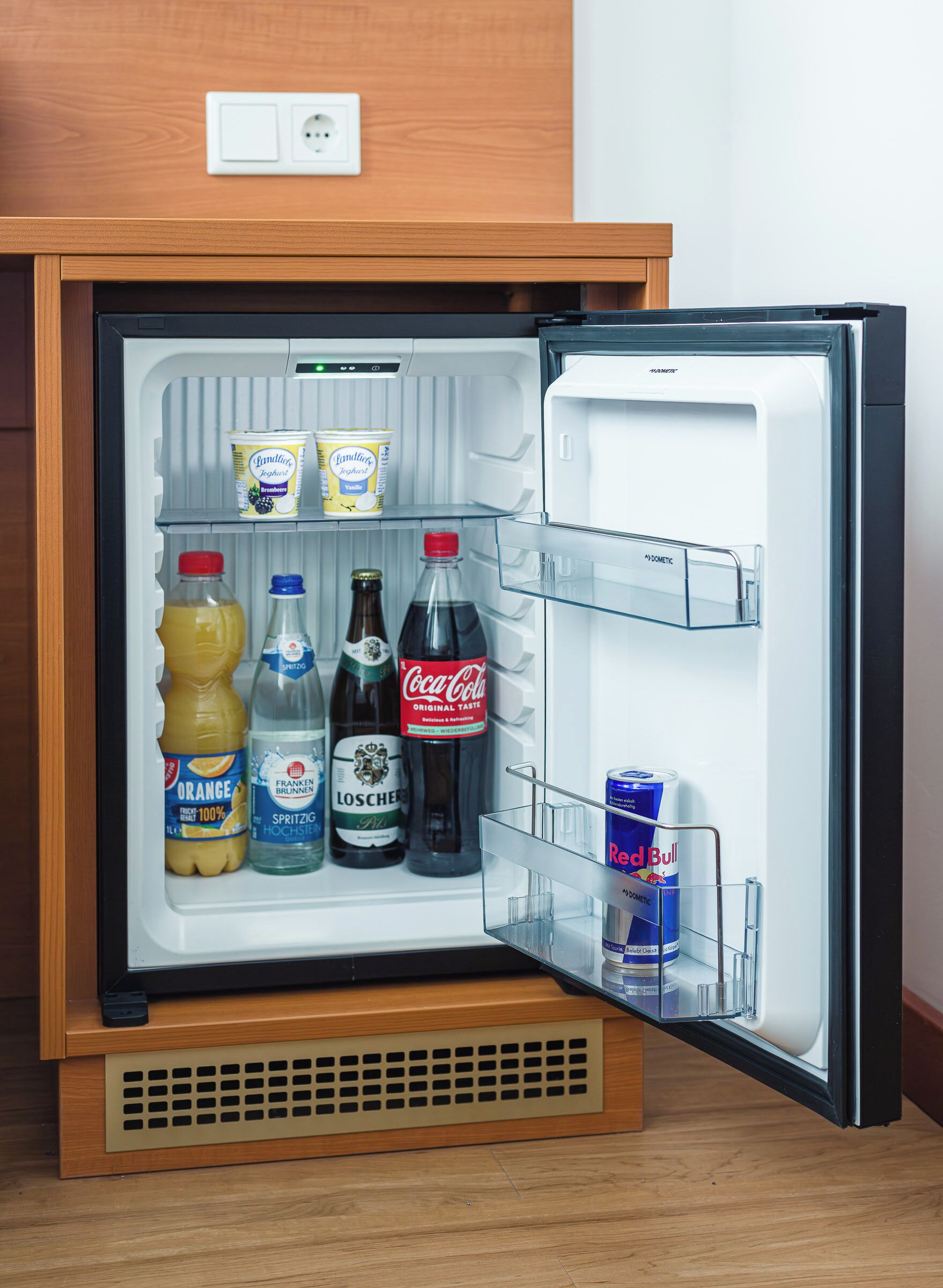 Un mini-réfrigérateur à l’extérieur en bois, contenant des boissons comme du jus d’orange, de la bière et des sodas.