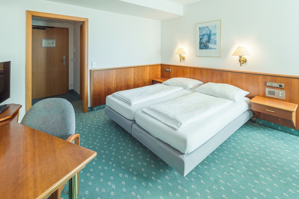 A hotel room with a double bed, a wooden desk, a chair, two bedside tables, and a wall-mounted lamp.