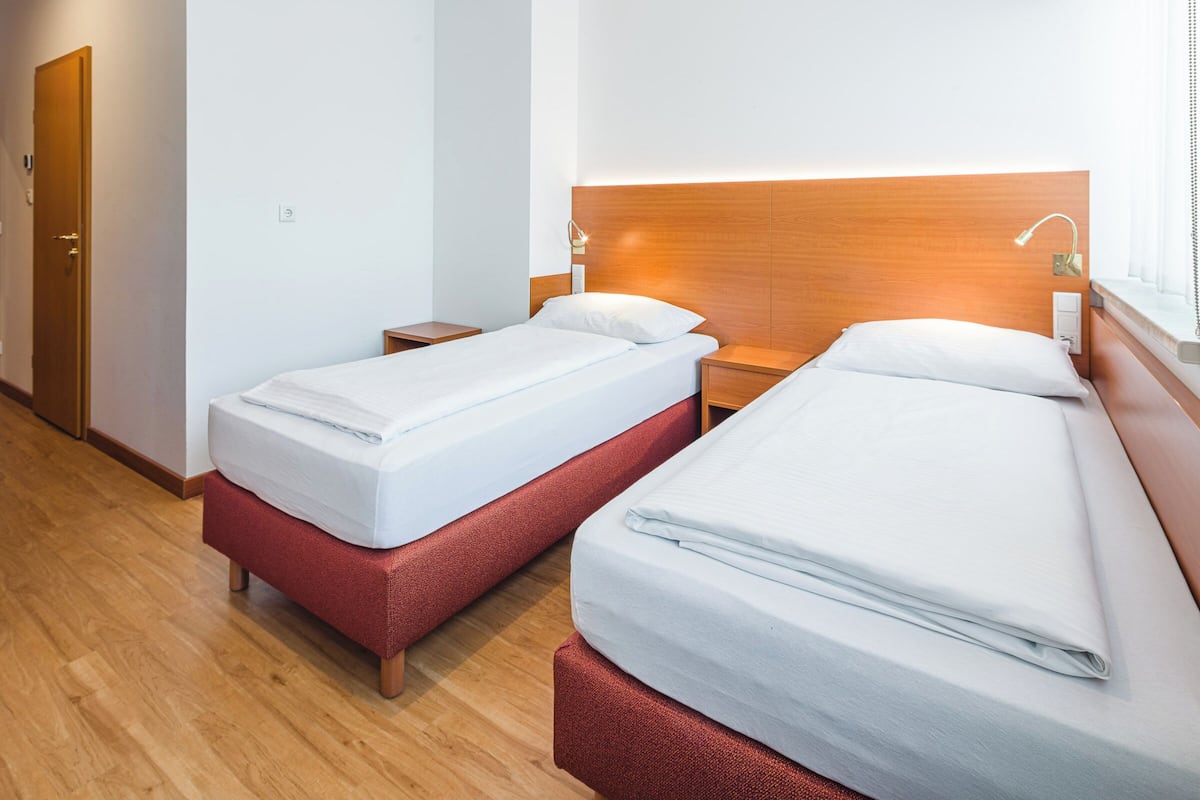 A hotel room with two single beds, wooden headboards, bedside tables with lamps, and a wooden floor.