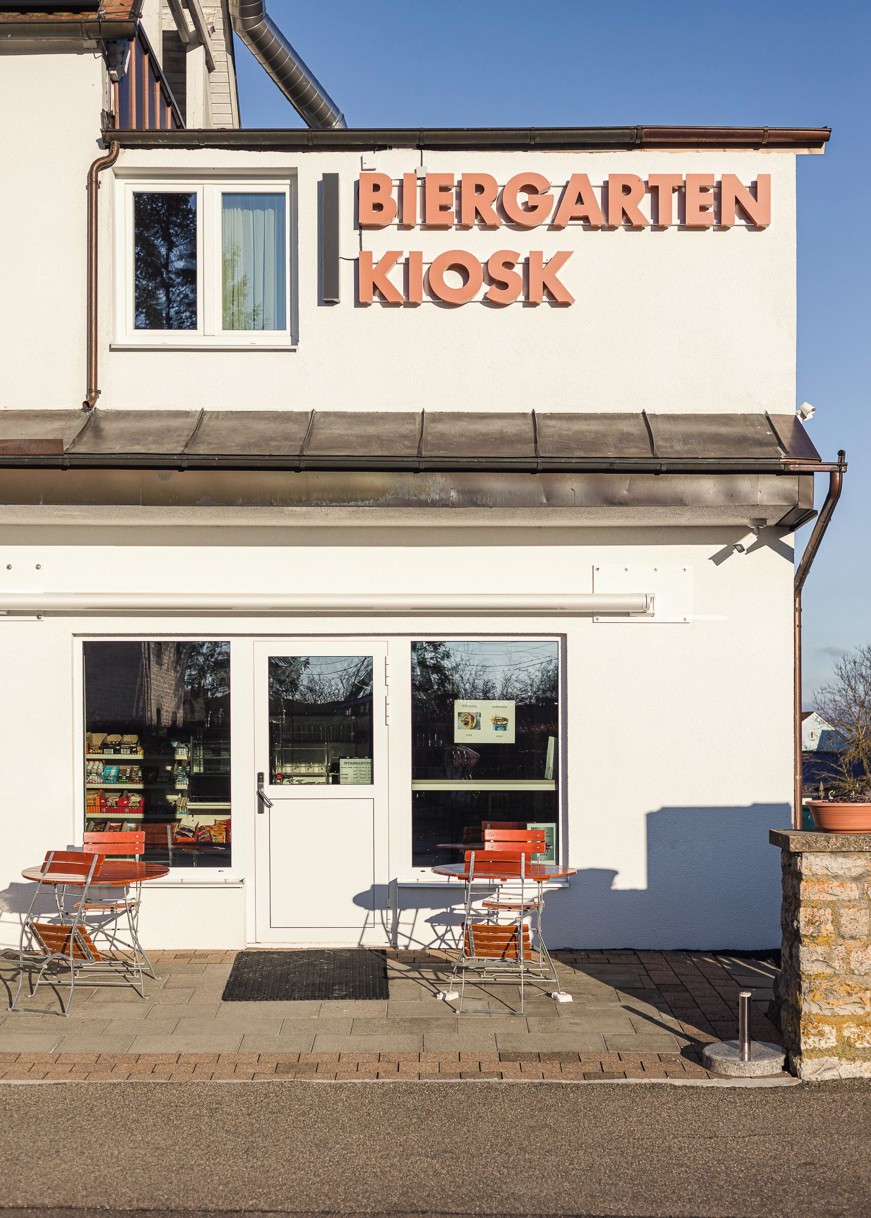Ein Gebäude mit einem Schild, auf dem "BIERGARTEN KIOSK" steht, und Außenbestuhlung mit Stühlen und Tischen.
