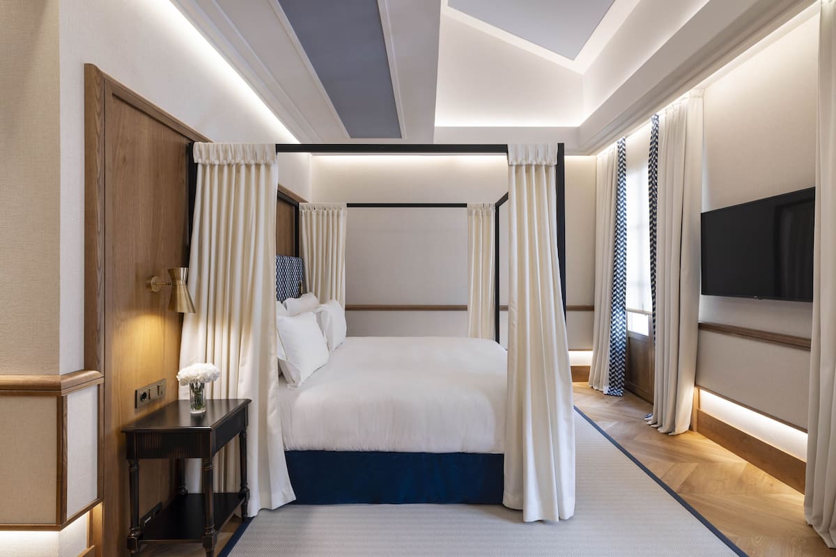 A modern hotel room with a large bed, a bedside table with a lamp, and a flat-screen TV mounted on the wall.