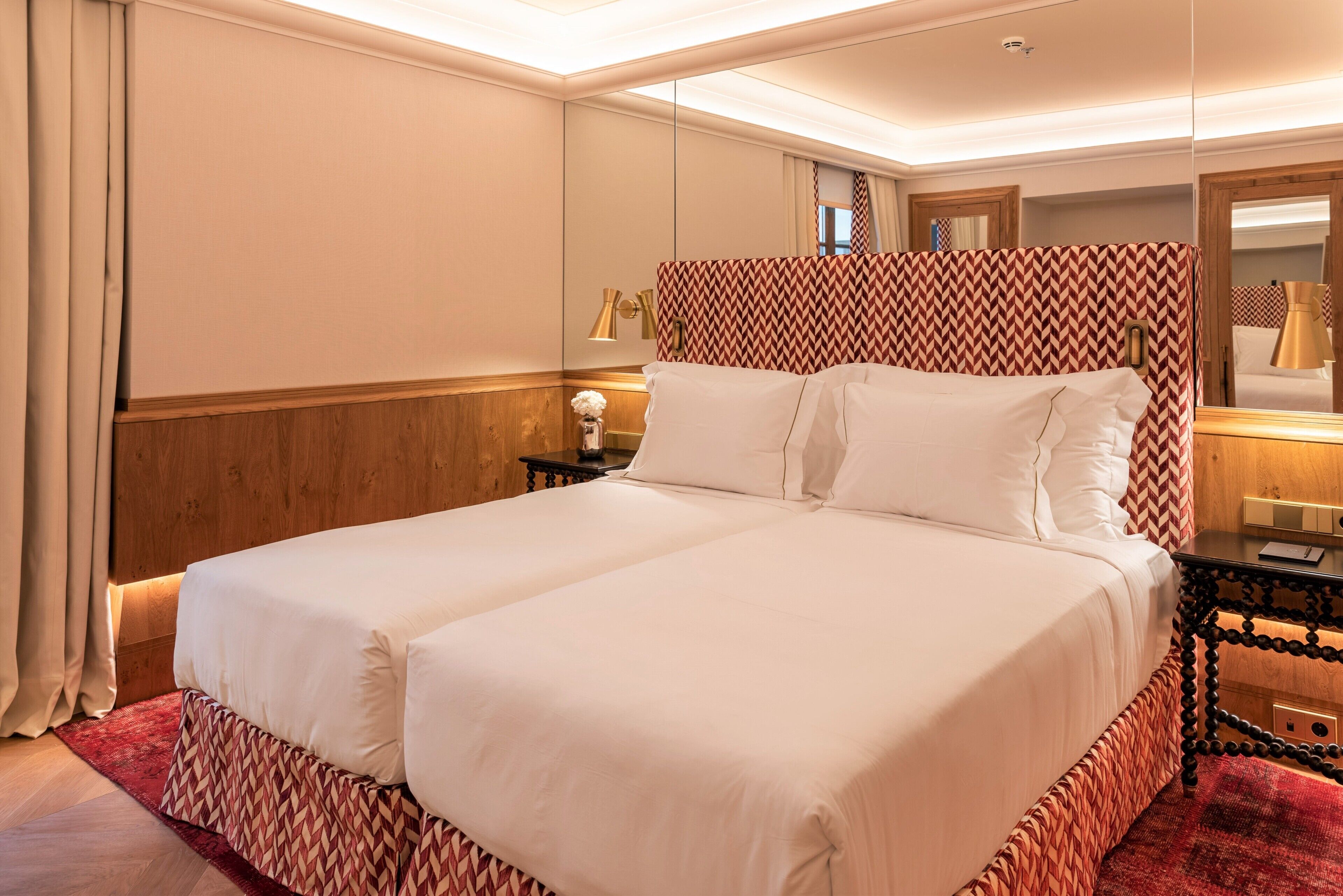 Una cama doble con cabecera estampada, una mesita de noche con lámpara y una alfombra roja.