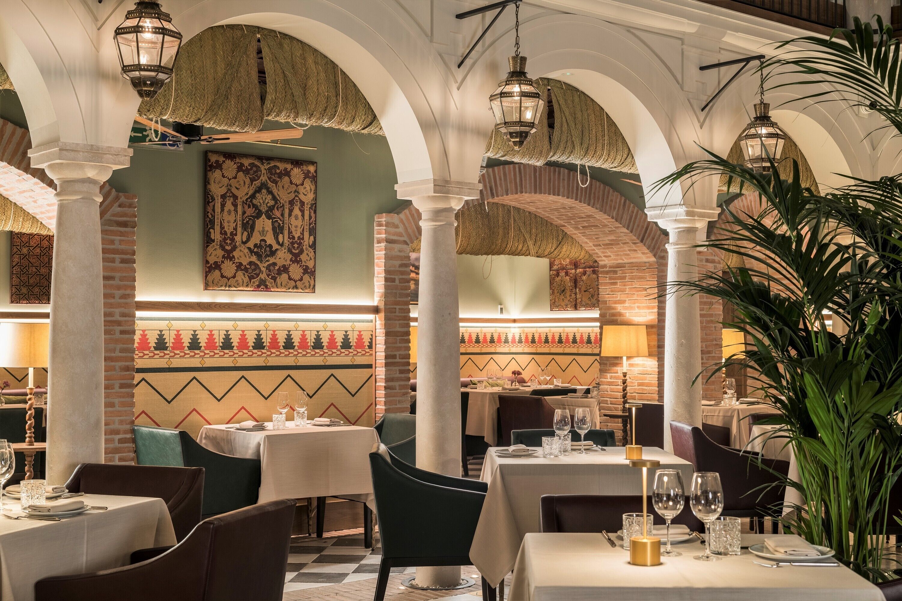 Un restaurante con techos altos en arco, paredes de ladrillo visto y lámparas tradicionales. Las mesas están dispuestas con manteles blancos y copas de vino.