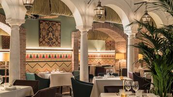 Un restaurante con techos altos en arco, paredes de ladrillo visto y lámparas tradicionales. Las mesas están dispuestas con manteles blancos y copas de vino.