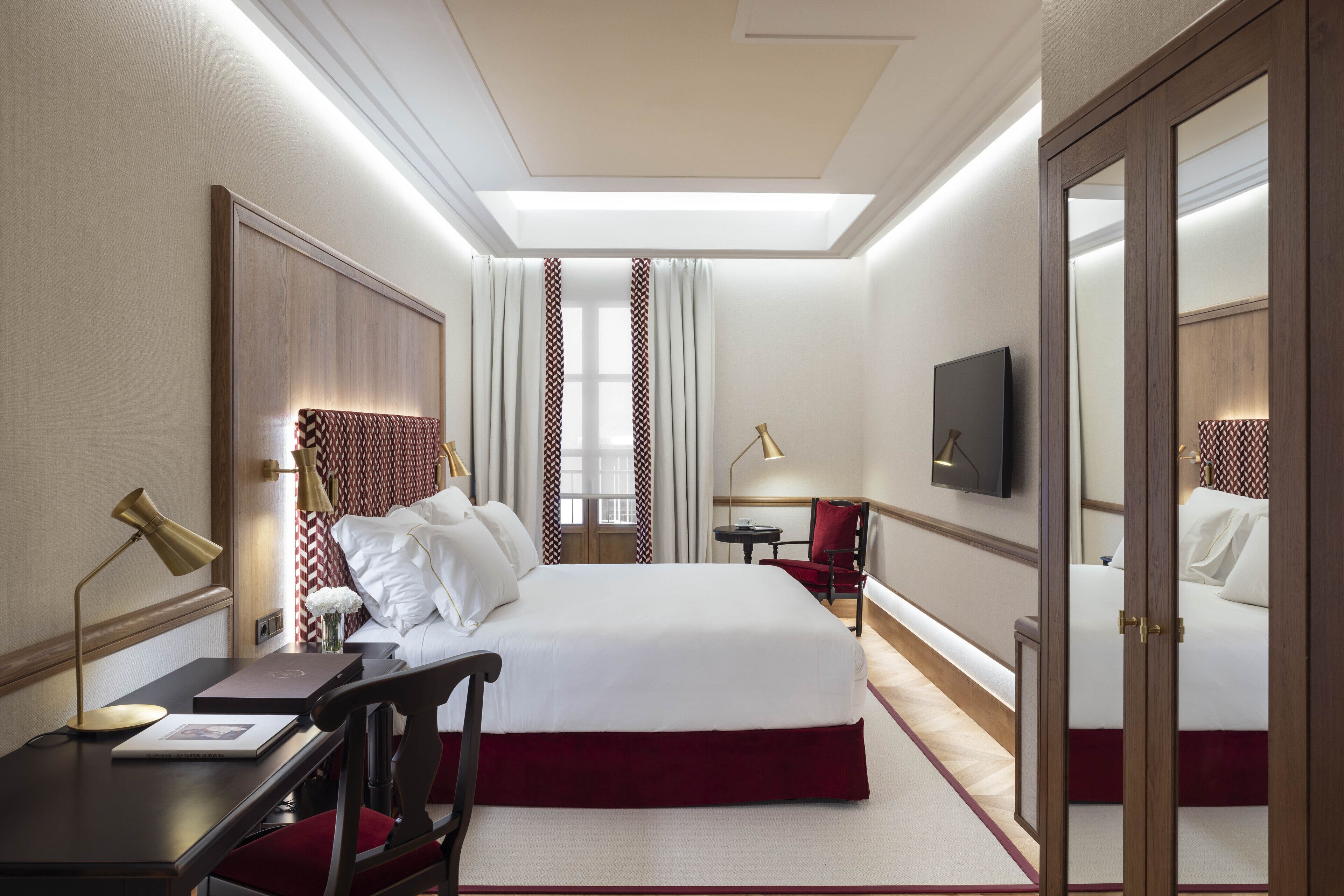 A hotel room with a large bed, bedside tables, a desk, a chair, a TV, and a mirror.