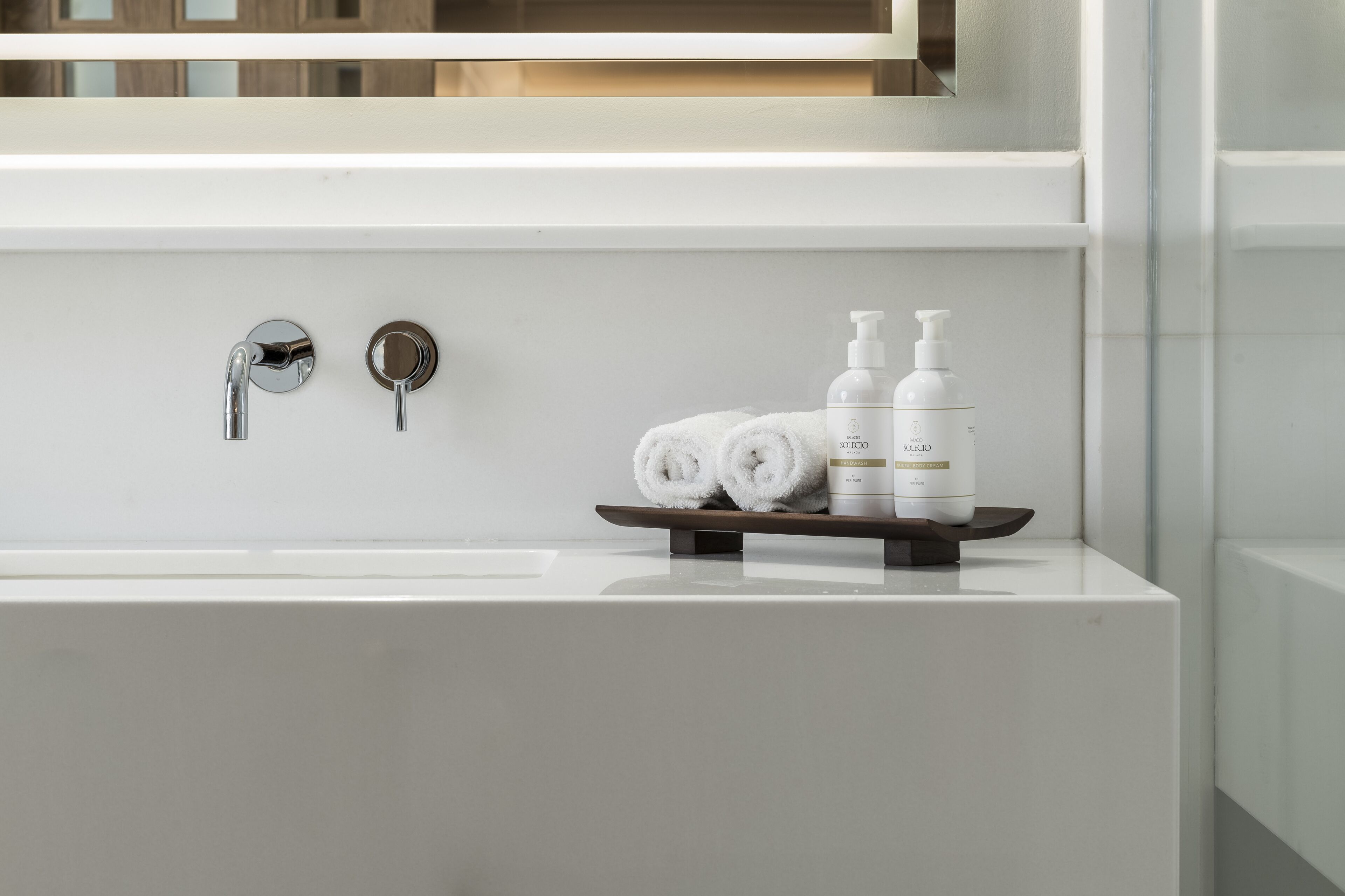 A modern bathroom with a white sink, two faucets, and a shelf holding rolled towels and toiletries.