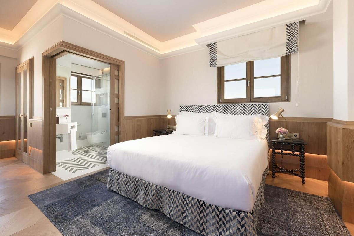 A hotel room with a large bed, a bedside table with a lamp, a mirror, and a window with patterned curtains.