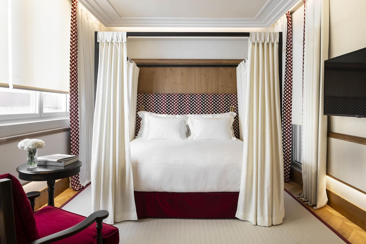 A neatly arranged bedroom with a bed, curtains, a TV, a small table with a vase, and a chair.