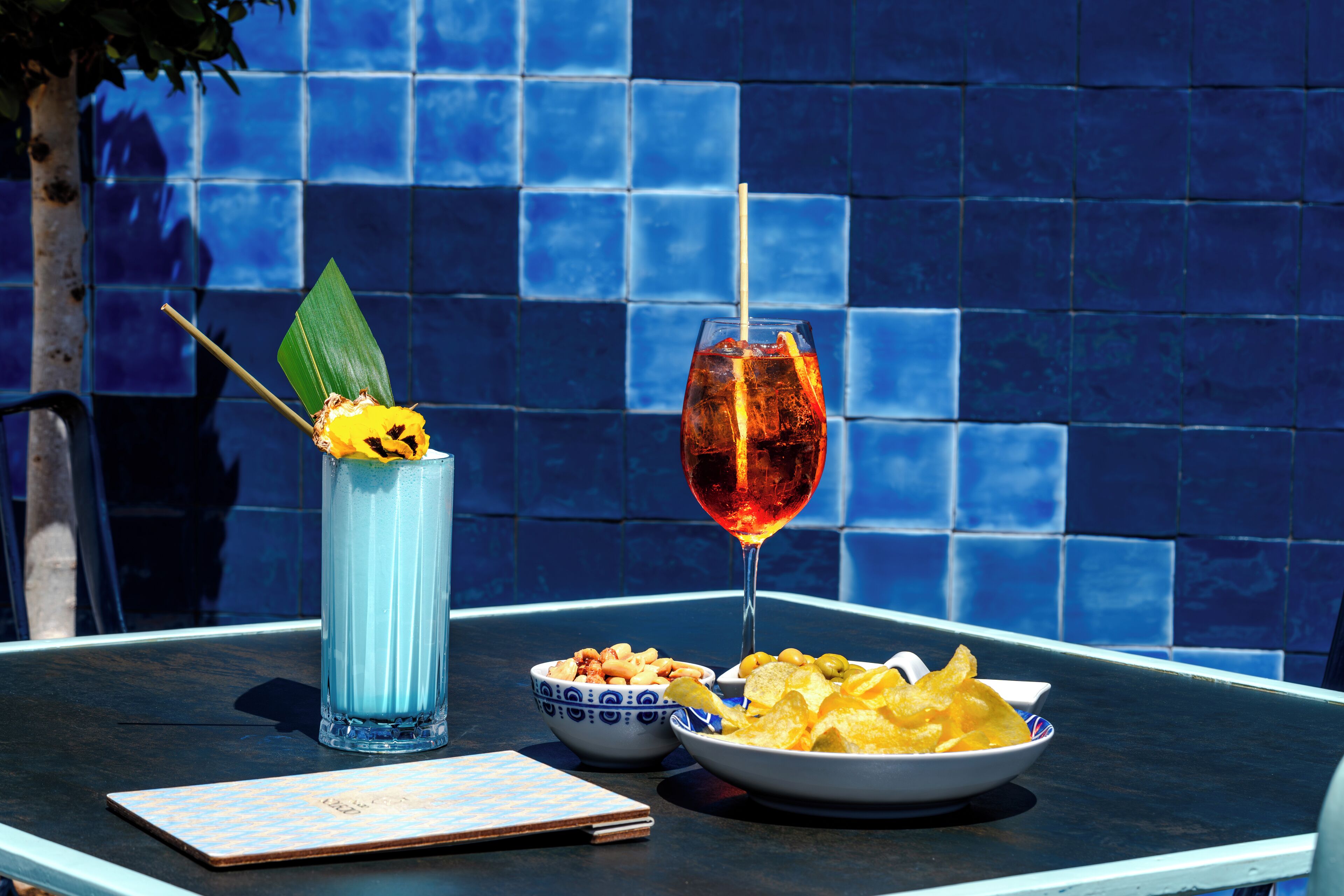 A table with a blue tiled wall, a glass of red drink, a bowl of chips, and a menu.