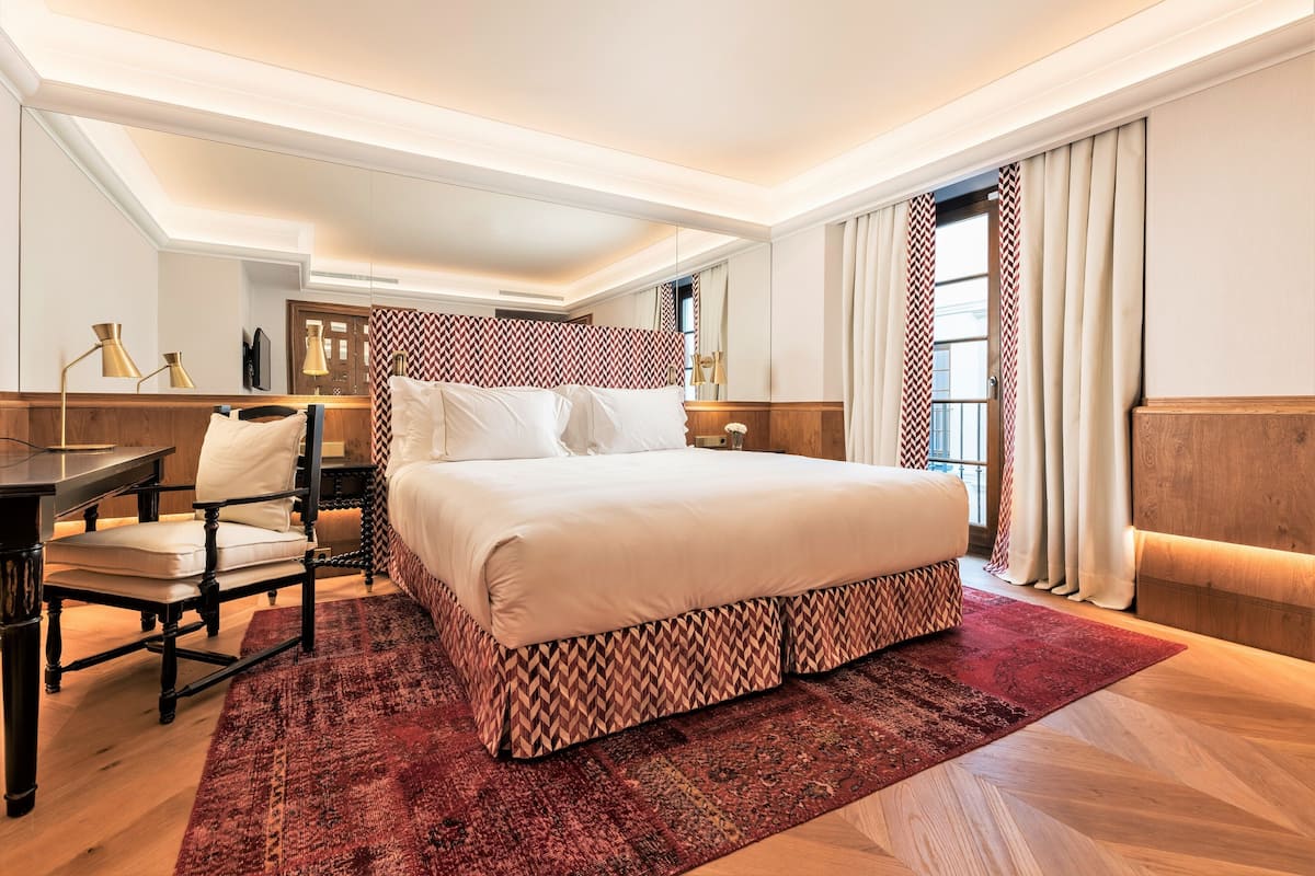 A modern hotel room with a large bed, a desk, and a chair. The room has a patterned rug and a large window with curtains.