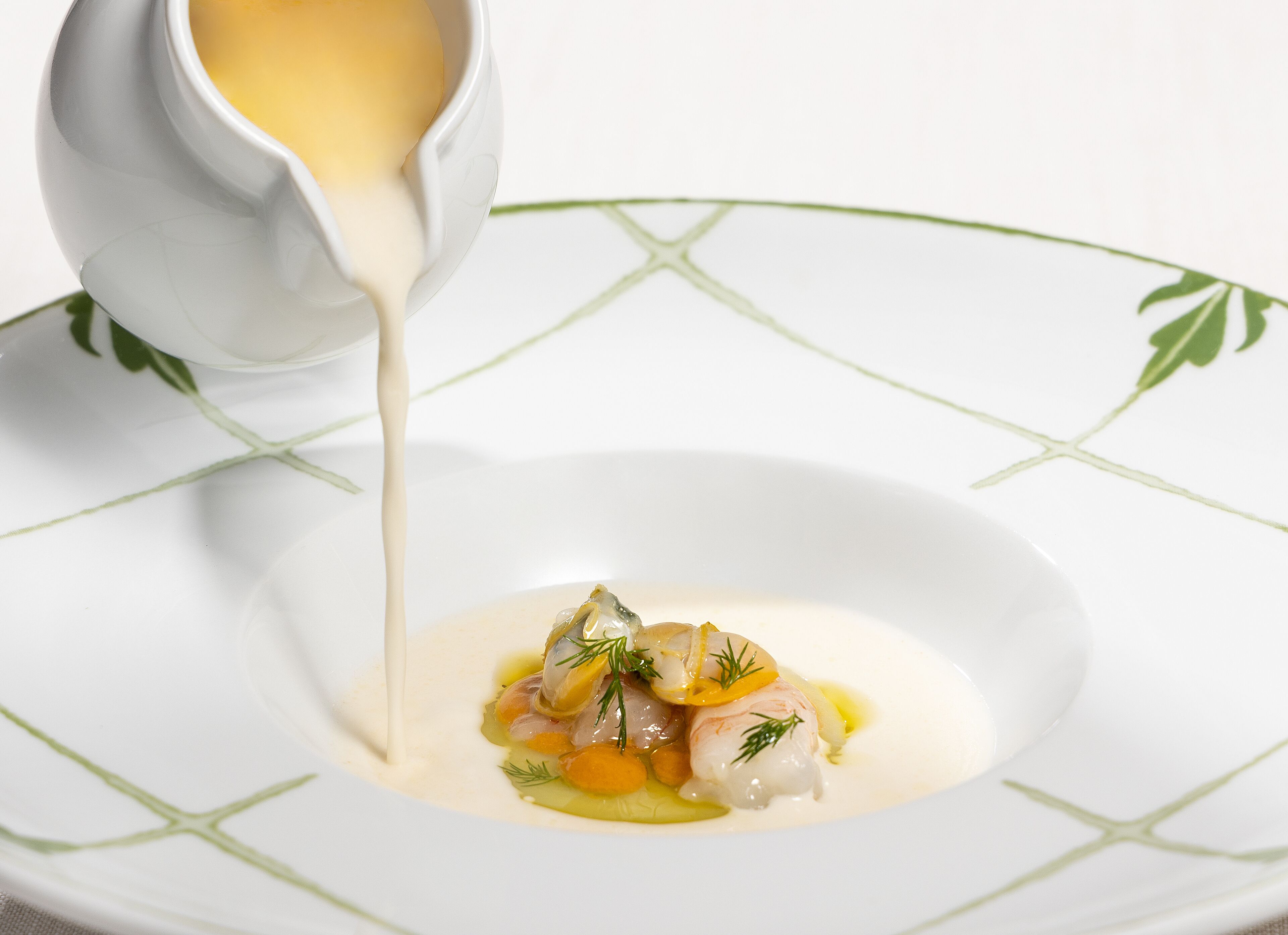 Une assiette blanche ornée d’un motif de feuilles vertes, contenant un plat composé de poisson, de légumes et d’une sauce crémeuse versée à partir d’un pichet.