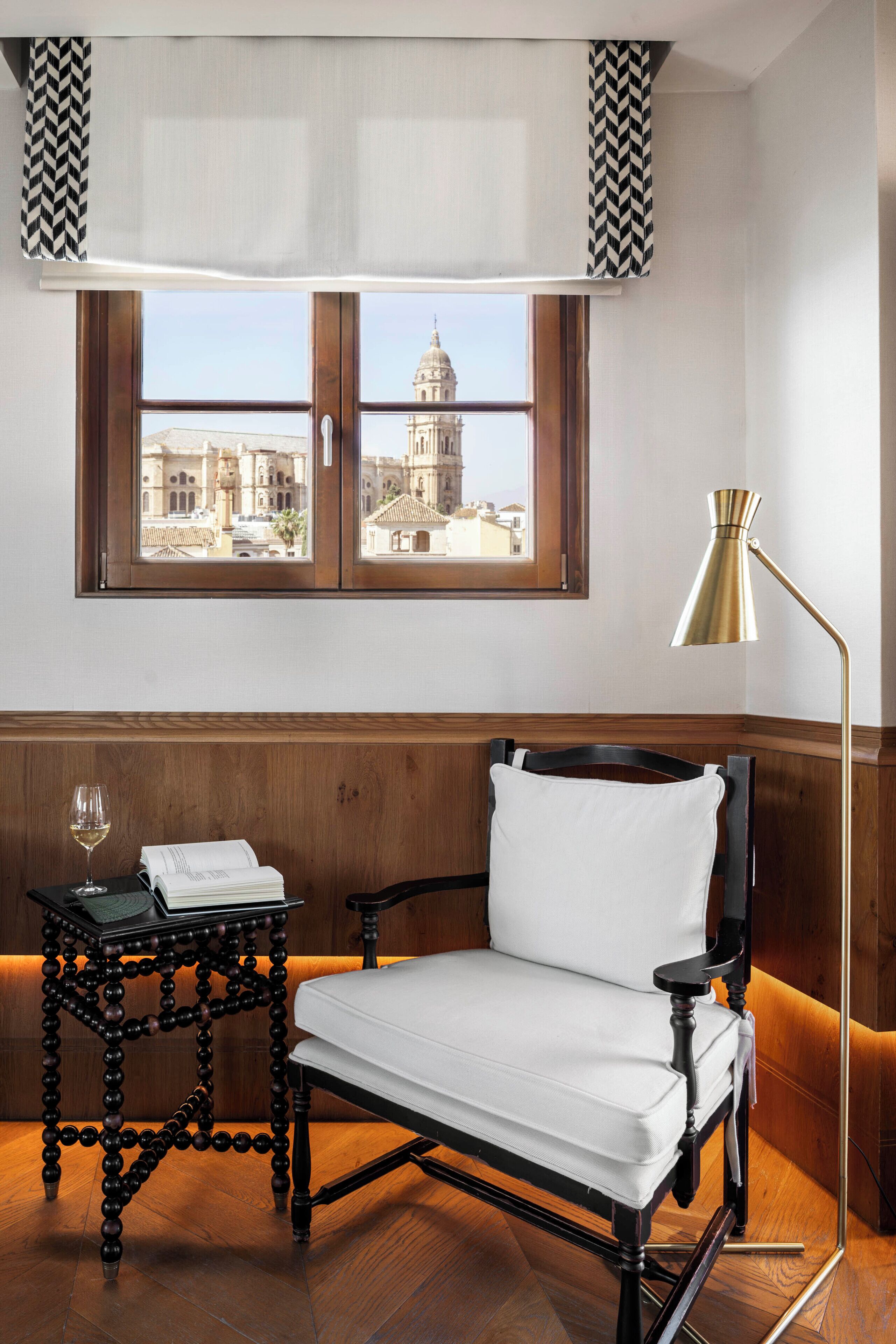 Une pièce avec vue sur un bâtiment historique, visible à travers une fenêtre, comprenant une chaise en bois, une petite table avec un verre de vin et un lampadaire.