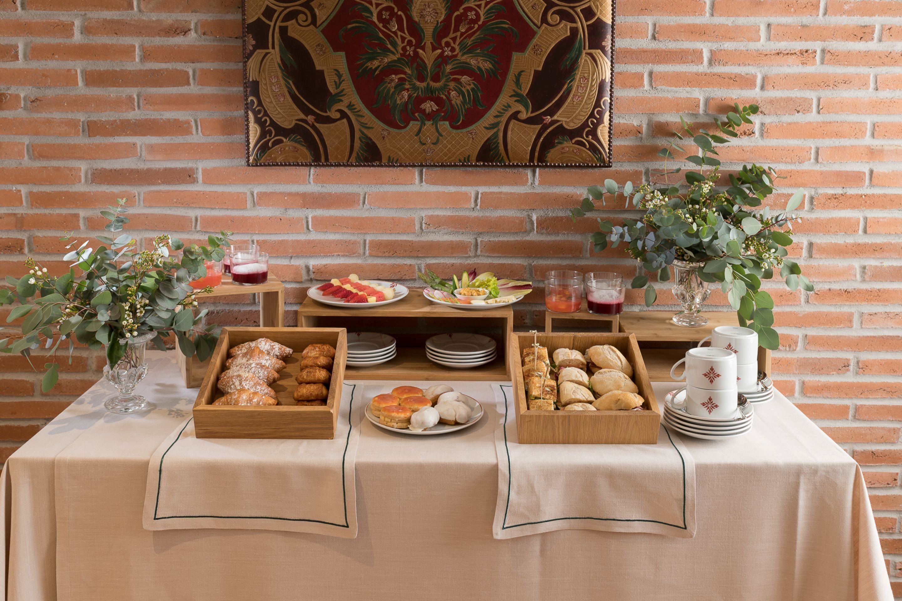 Una mesa de buffet con diversos alimentos y bebidas, frente a una pared de ladrillo con un tapiz decorativo.
