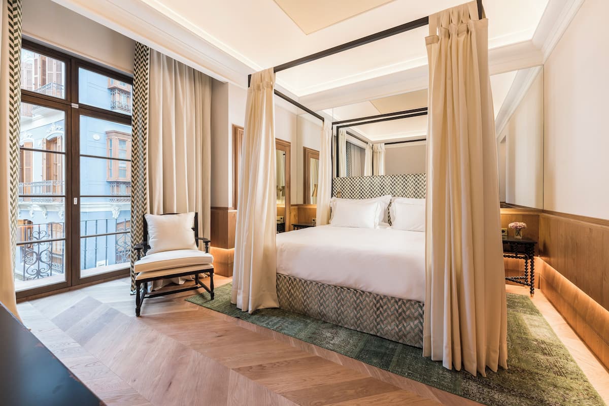 A hotel room with a large bed, a chair, a view of the outside balcony, and a herringbone-patterned floor.