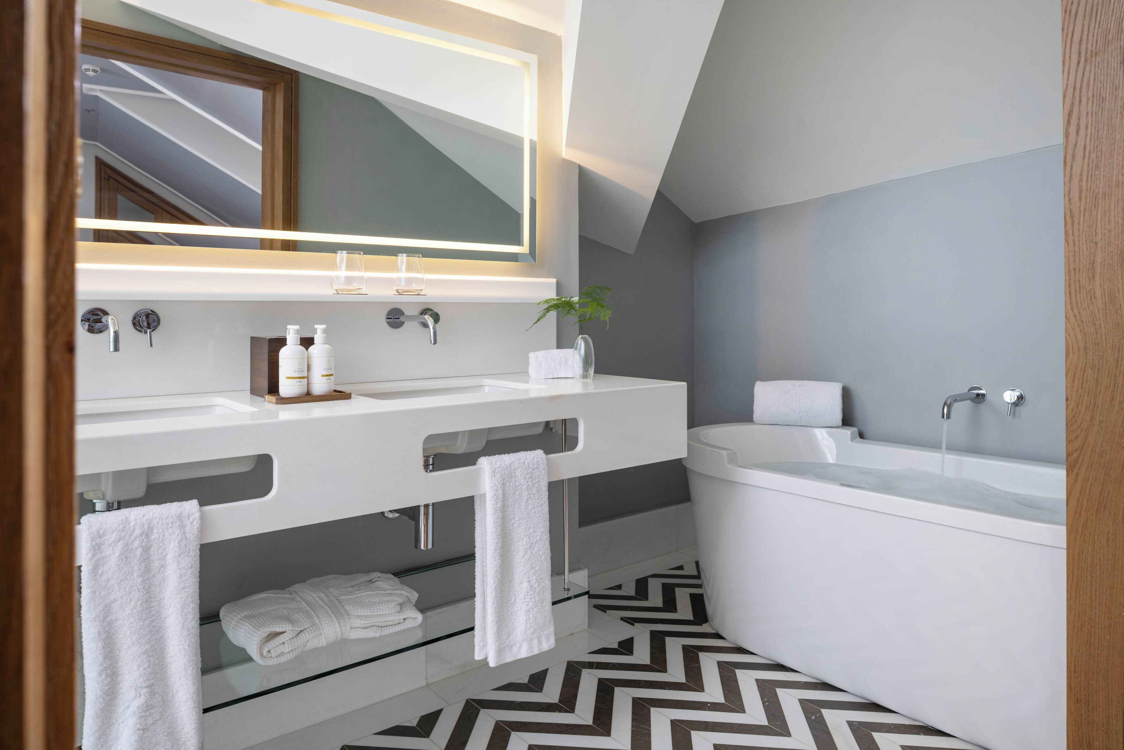 Un baño moderno con dos lavamanos, un espejo grande, una bañera y un suelo con estampado tipo espiga.