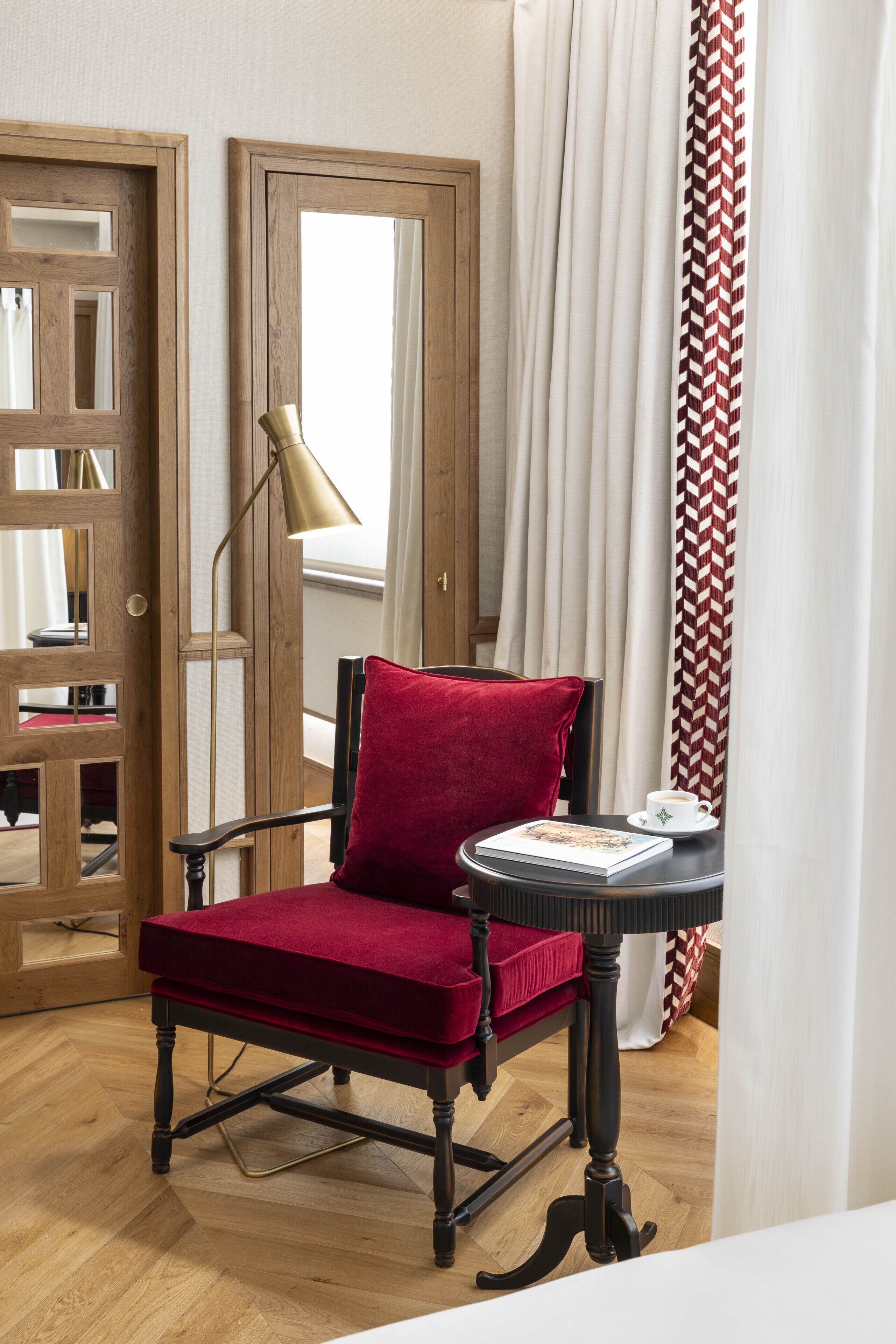 A red velvet chair with a black frame, a small round table with a book and a cup, and a floor lamp.