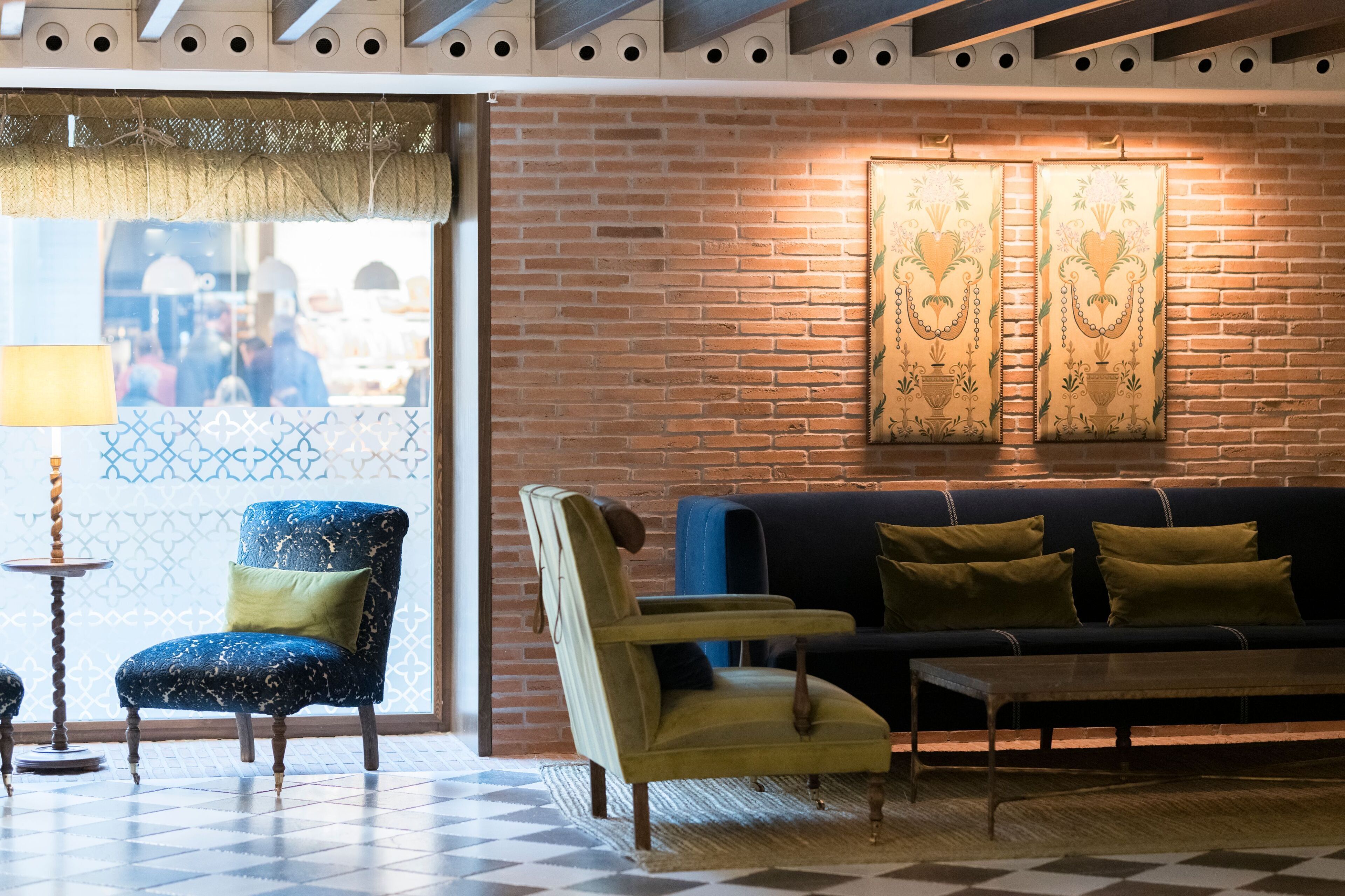 Una habitación con piso de cuadros, un sofá, dos sillones, una lámpara y una pared de ladrillo con cortinas estampadas.