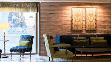 Una habitación con piso de cuadros, un sofá, dos sillones, una lámpara y una pared de ladrillo con cortinas estampadas.