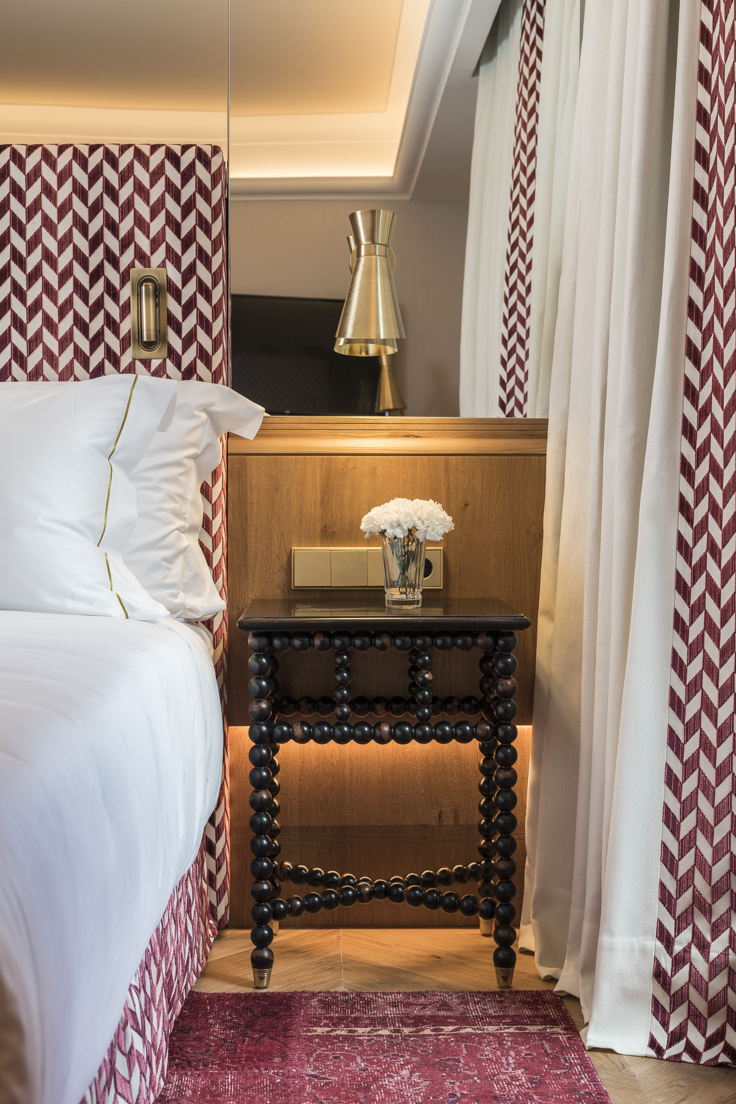 A neatly made bed with white linens, a wooden nightstand with a vase of flowers, and a patterned rug.