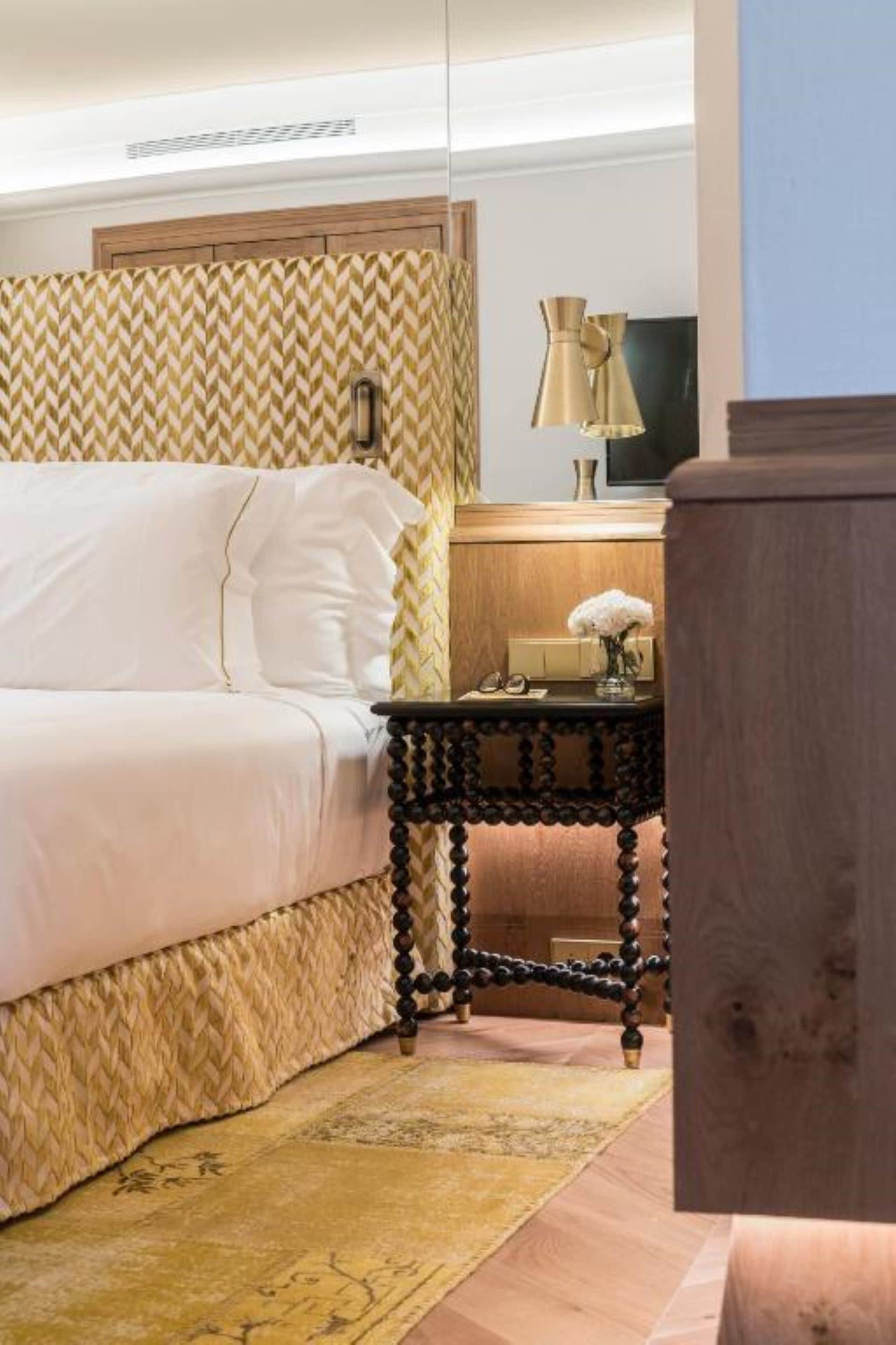 Un dormitorio con una cama, una mesita de noche de madera, una lámpara y un televisor instalado en la pared.