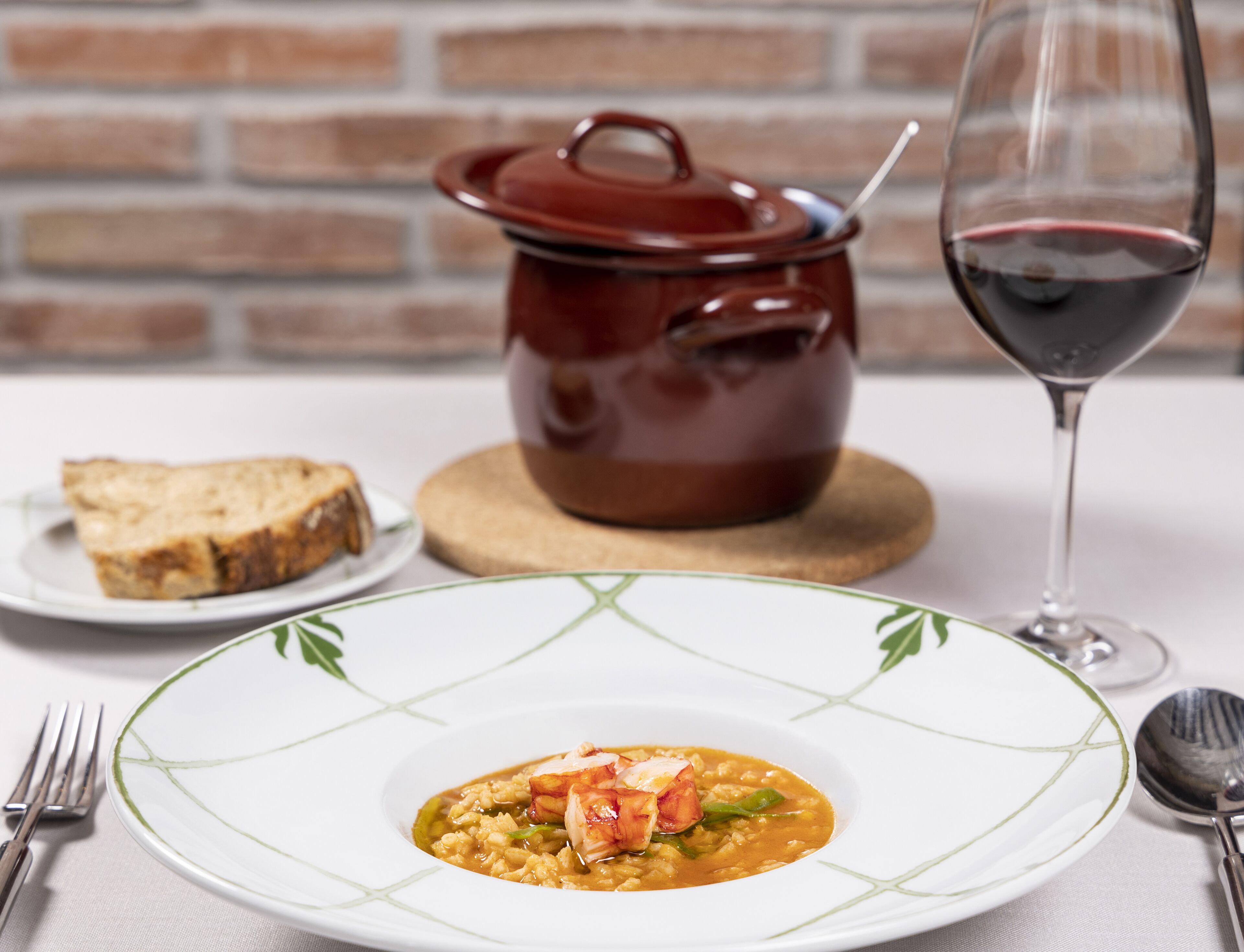 Una mesa de comedor con un plato de comida, una copa de vino tinto y una olla roja con tapa.