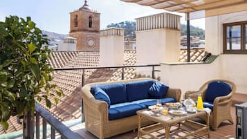 Un balcón con mobiliario de mimbre, una mesa con alimentos para el desayuno y una vista de una torre al fondo.