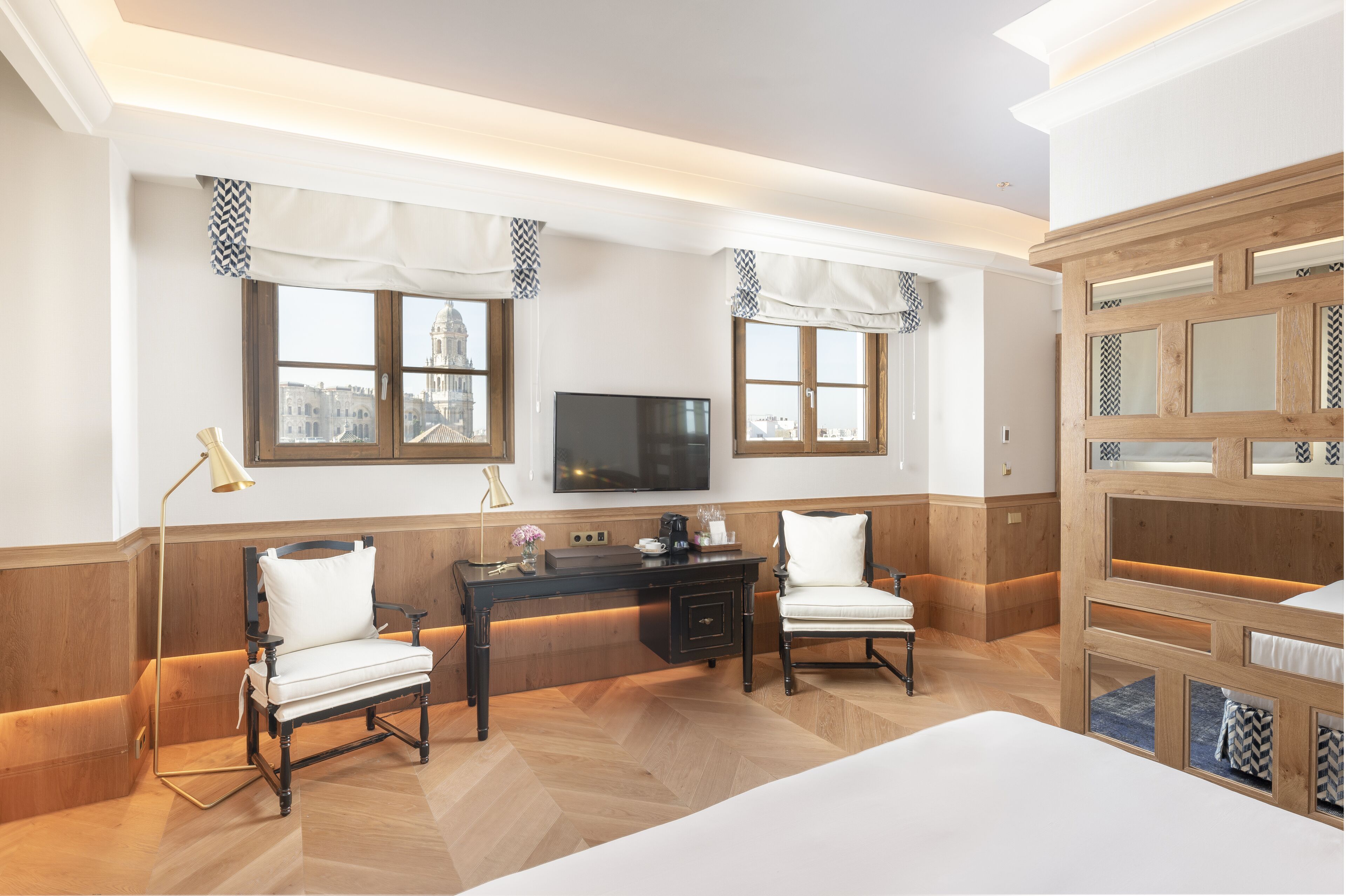 Quarto de hotel com escrivaninha de madeira, duas cadeiras, uma cama e uma televisão.