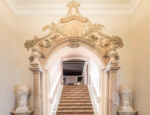 A grand staircase with ornate carvings and a patterned carpet, leading to a room with a high ceiling and decorative moldings.