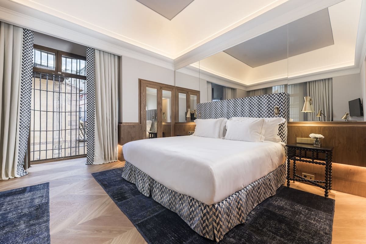 A modern hotel room with a large bed, a wooden nightstand, a mirror, and a view of the city through large windows.