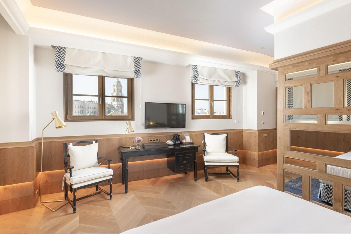 A hotel room with a wooden floor, a desk with a chair, a bed, a TV, and windows with curtains.